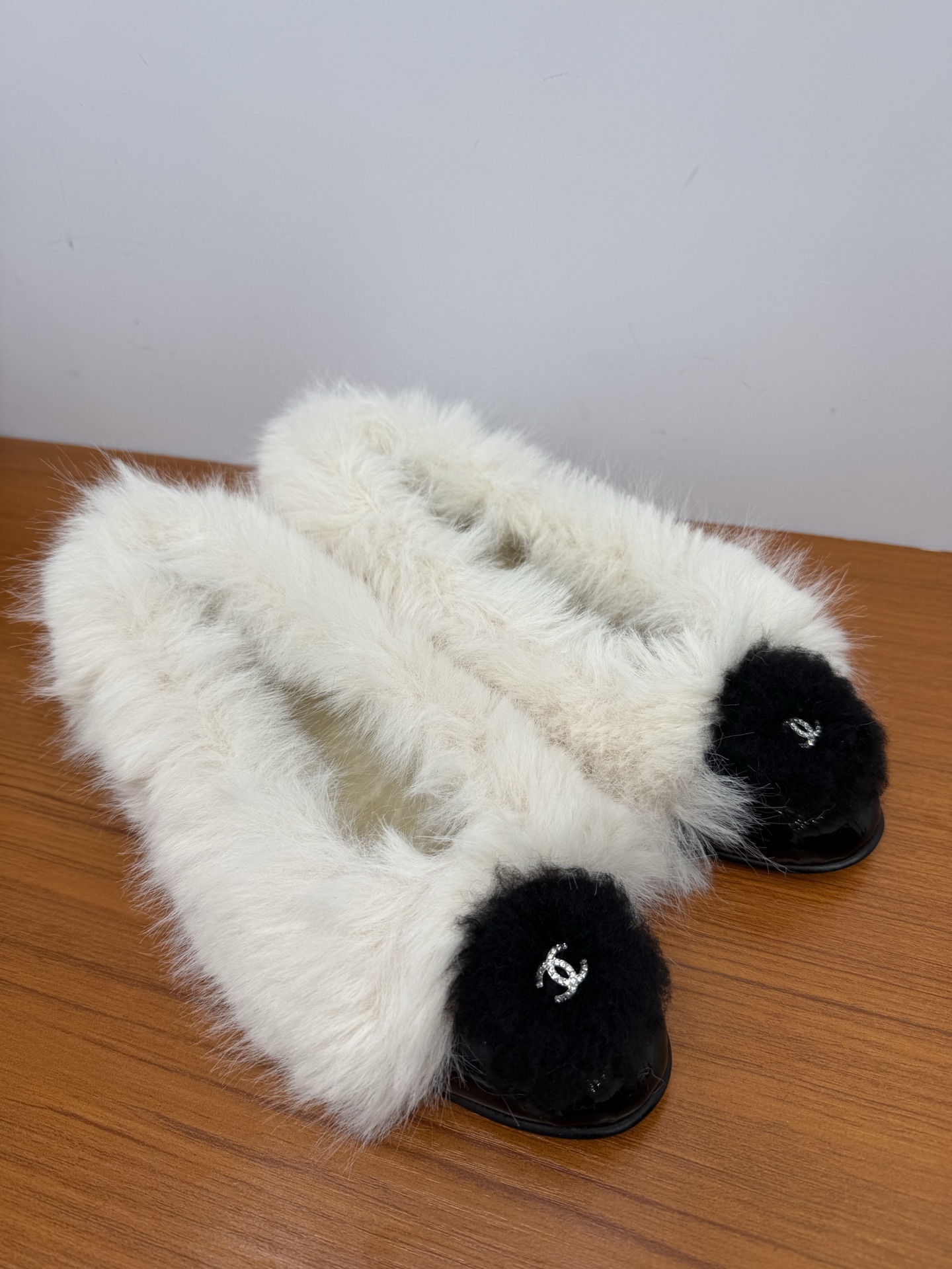Pair of white faux fur slippers with black fur toe caps and silver crystal CC logos on a wooden surface - Hình 2