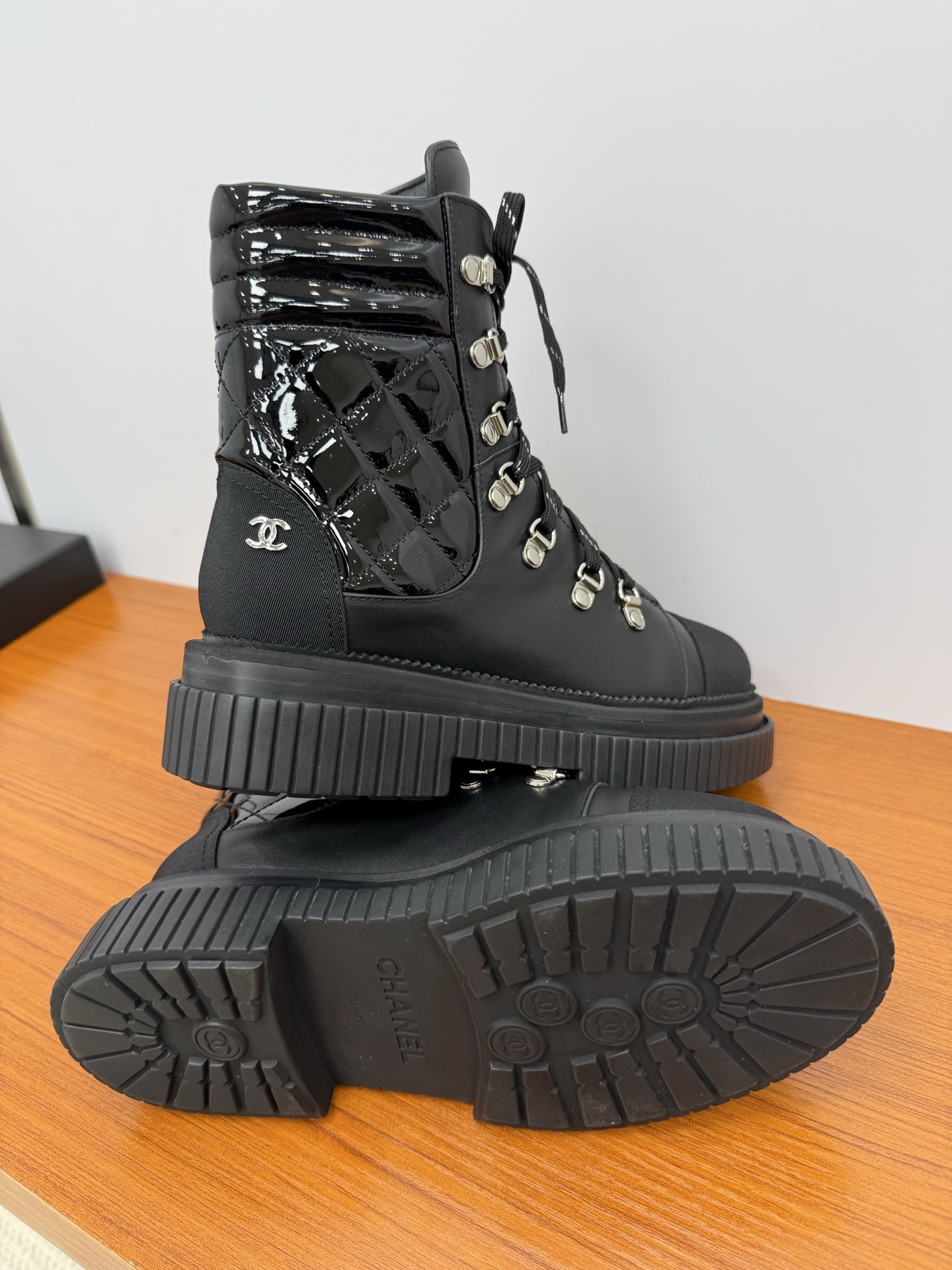 A pair of women's black leather lace-up combat boots with platform lug soles and quilted patent side panels on a wooden surface. - Hình 9