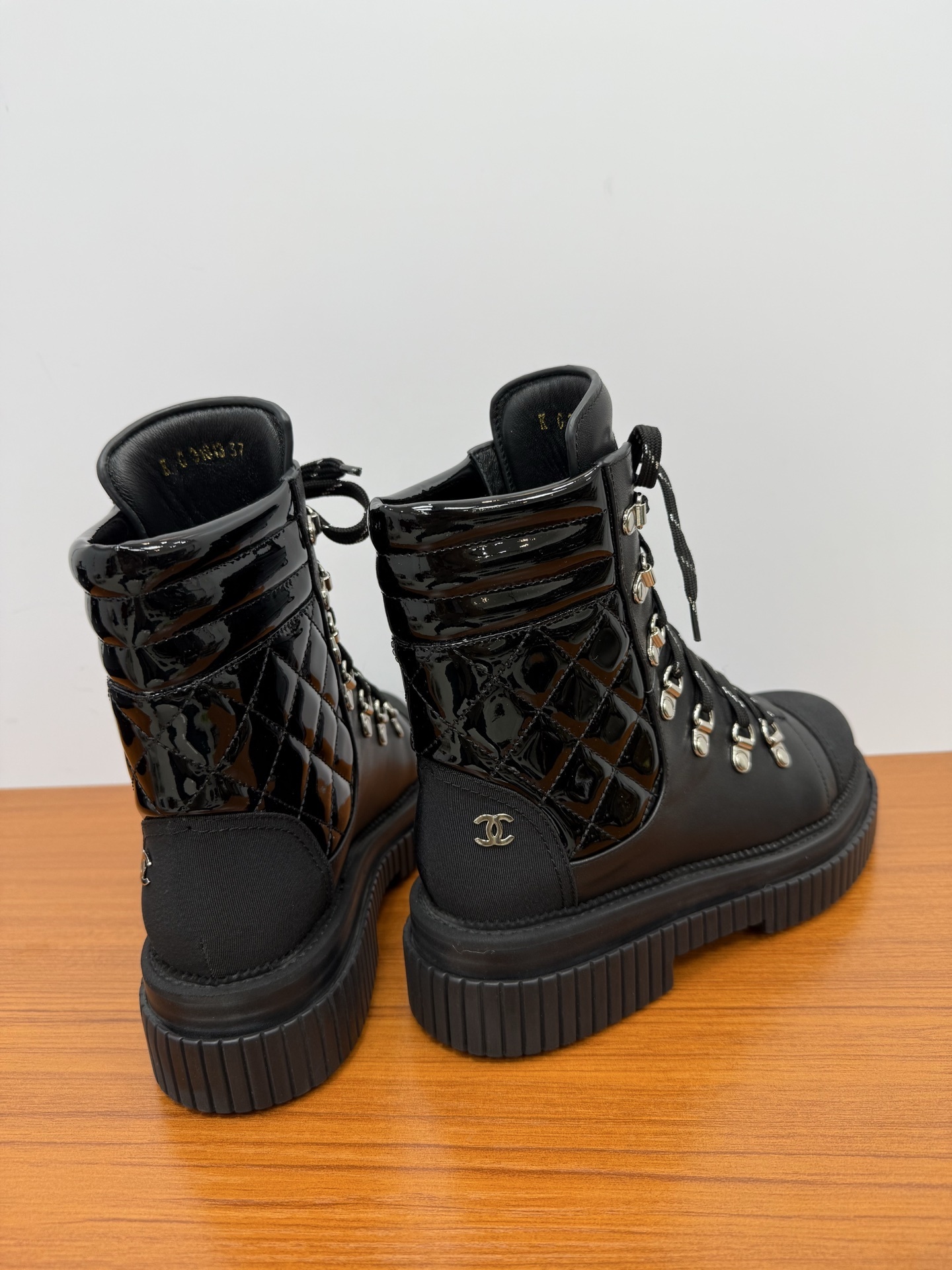 A pair of women's black leather lace-up combat boots with platform lug soles and quilted patent side panels on a wooden surface. - Hình 8