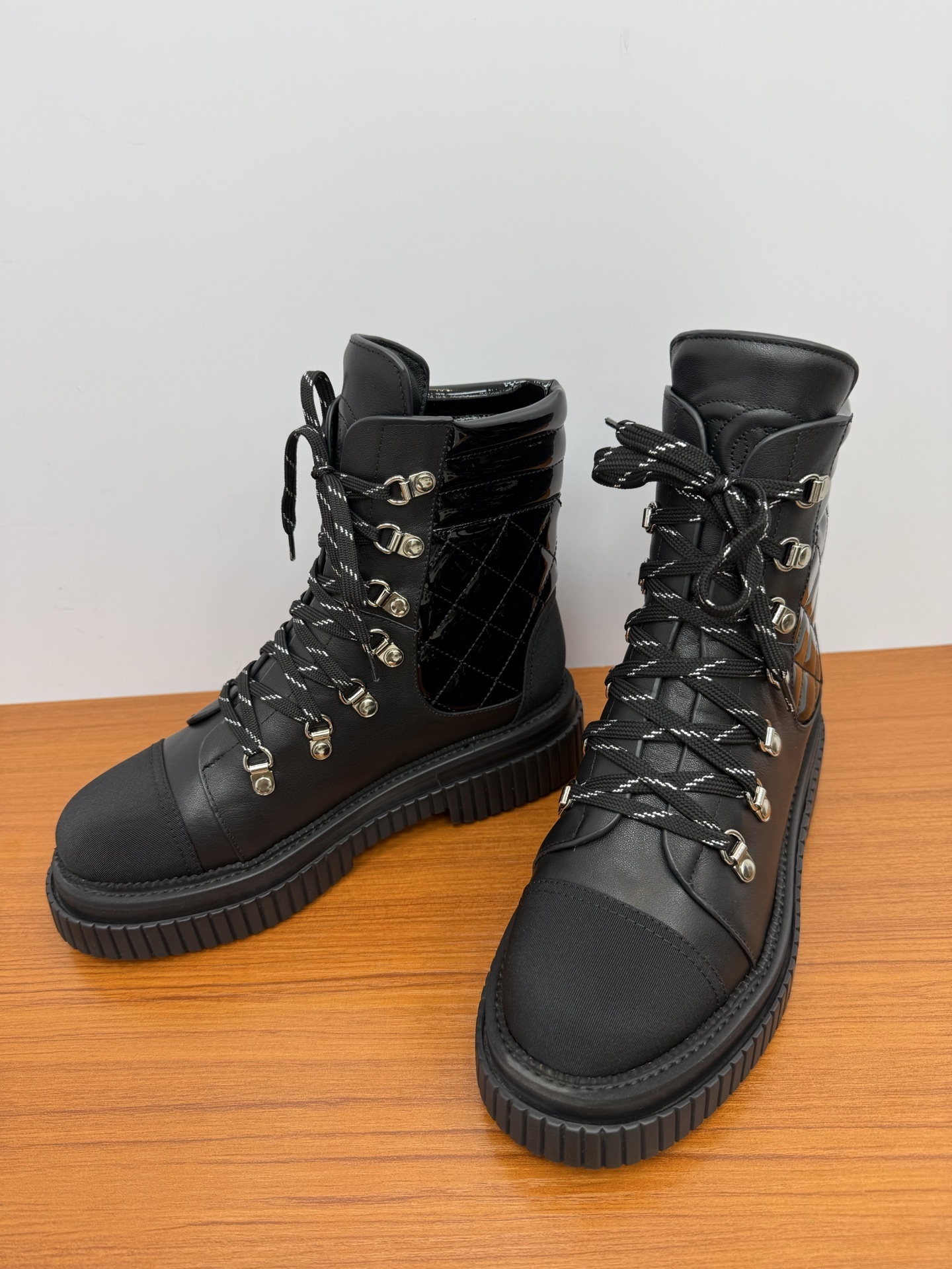 A pair of women's black leather lace-up combat boots with platform lug soles and quilted patent side panels on a wooden surface. - Hình 7