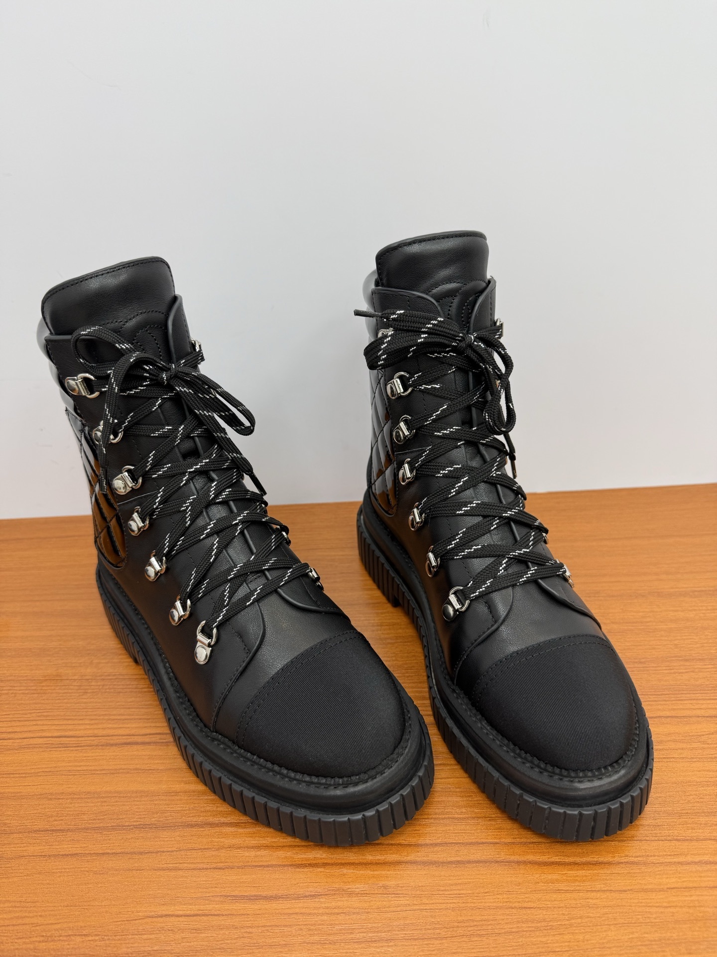 A pair of women's black leather lace-up combat boots with platform lug soles and quilted patent side panels on a wooden surface. - Hình 5