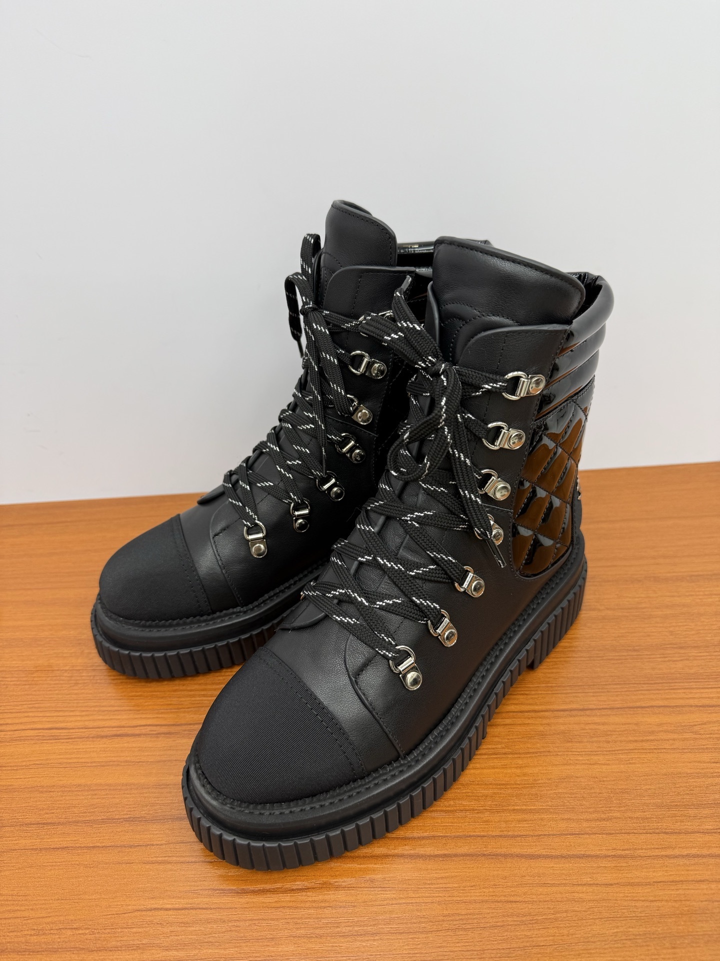 A pair of women's black leather lace-up combat boots with platform lug soles and quilted patent side panels on a wooden surface. - Hình 3