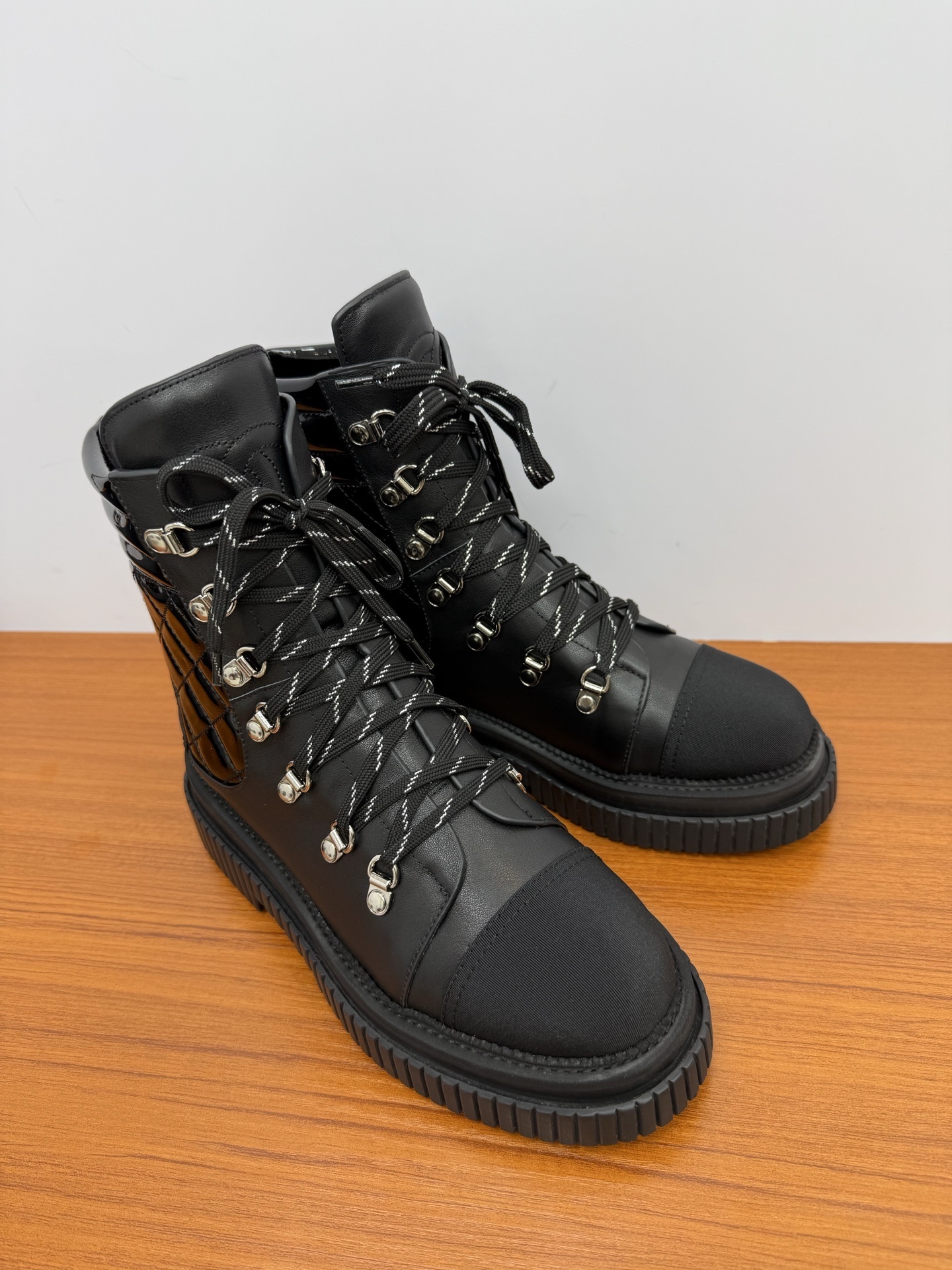 A pair of women's black leather lace-up combat boots with platform lug soles and quilted patent side panels on a wooden surface. - Hình 2