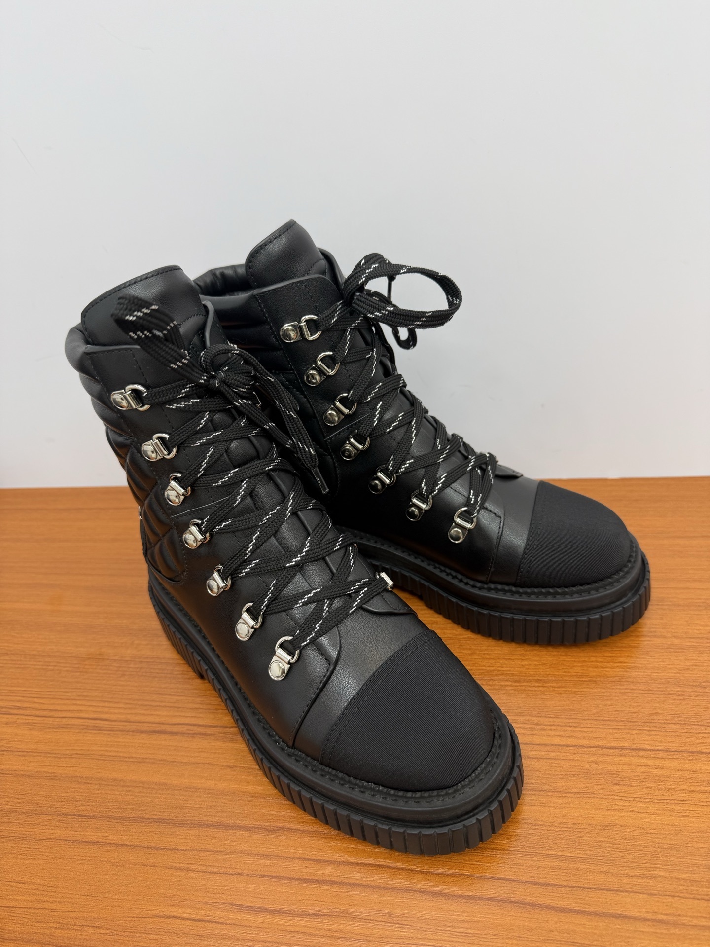 A pair of premium black quilted leather combat boots with chunky platform soles and patterned laces displayed on a wooden surface. - Hình 2