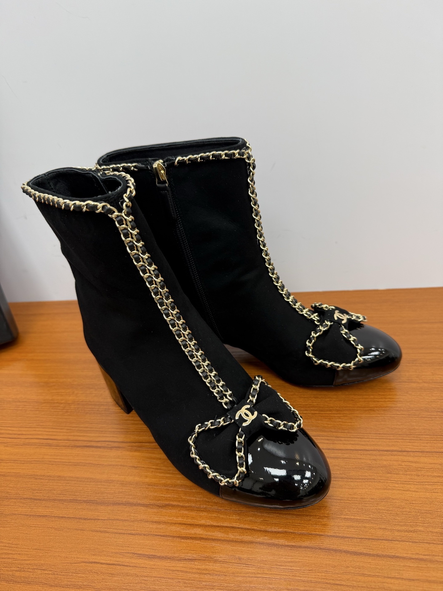 A pair of elegant black suede ankle boots with gold chain trim and a decorative bow on the patent leather toe cap. - Hình 2