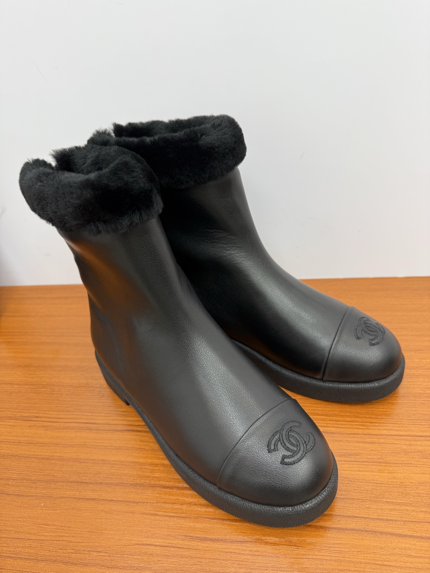 A pair of luxury Chanel black leather ankle boots featuring black fur trim at the collar and the embossed interlocking CC logo on the toe caps. - Hình 2