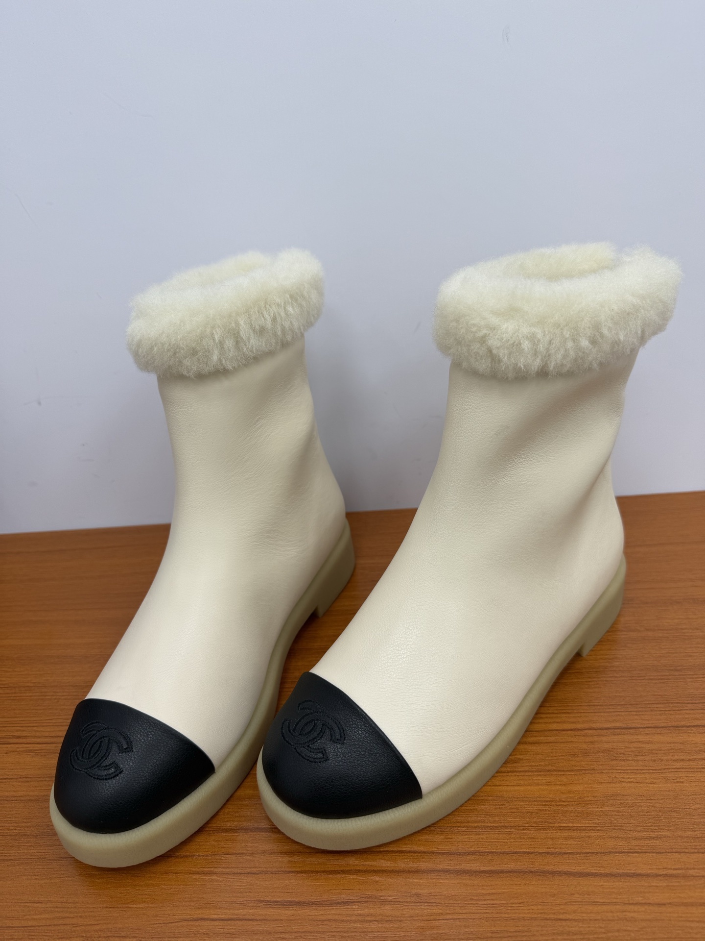 Pair of Chanel white leather ankle boots with black CC logo toe caps and white shearling trim on a wooden surface - Hình 6
