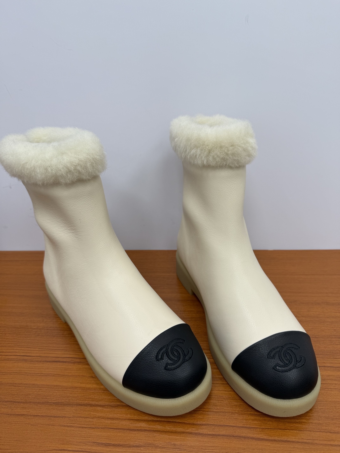 Pair of Chanel white leather ankle boots with black CC logo toe caps and white shearling trim on a wooden surface - Hình 5