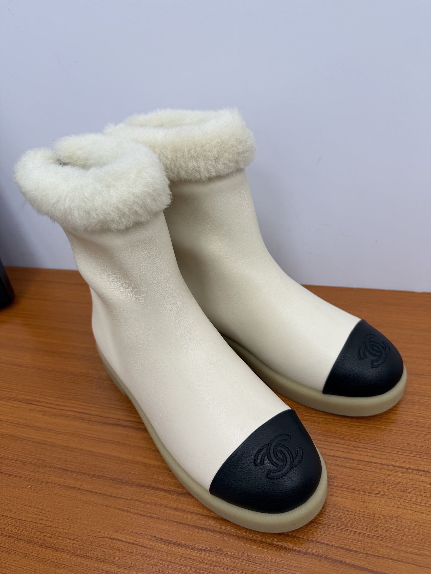 Pair of Chanel white leather ankle boots with black CC logo toe caps and white shearling trim on a wooden surface - Hình 2