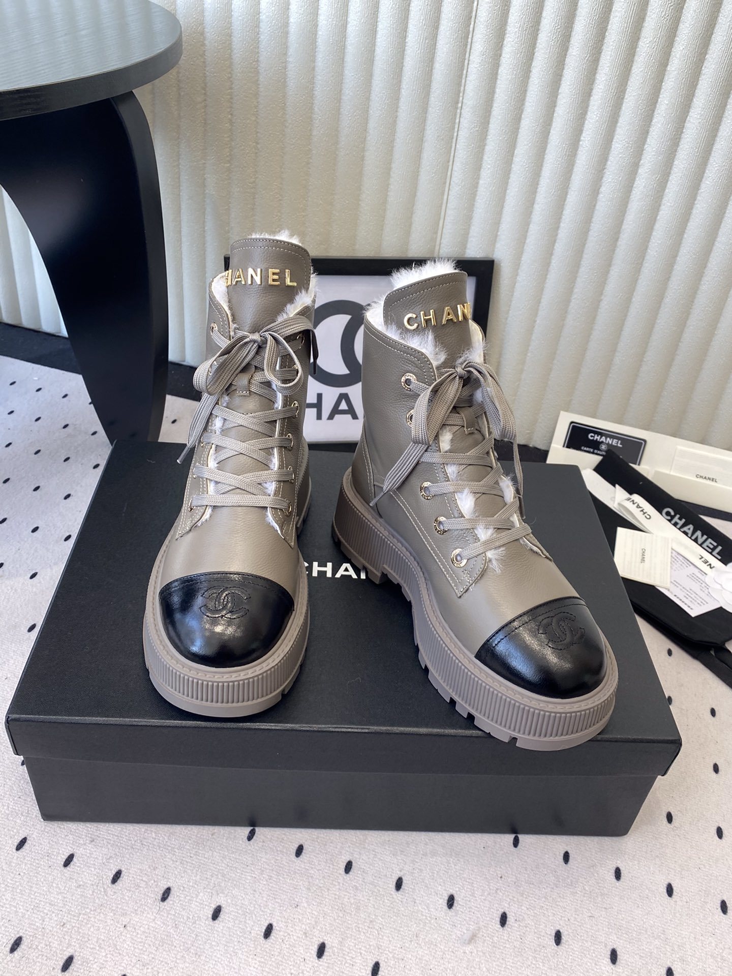 Chanel taupe leather lace-up boots with black patent toe caps and white shearling lining displayed on a black designer box - Hình 7