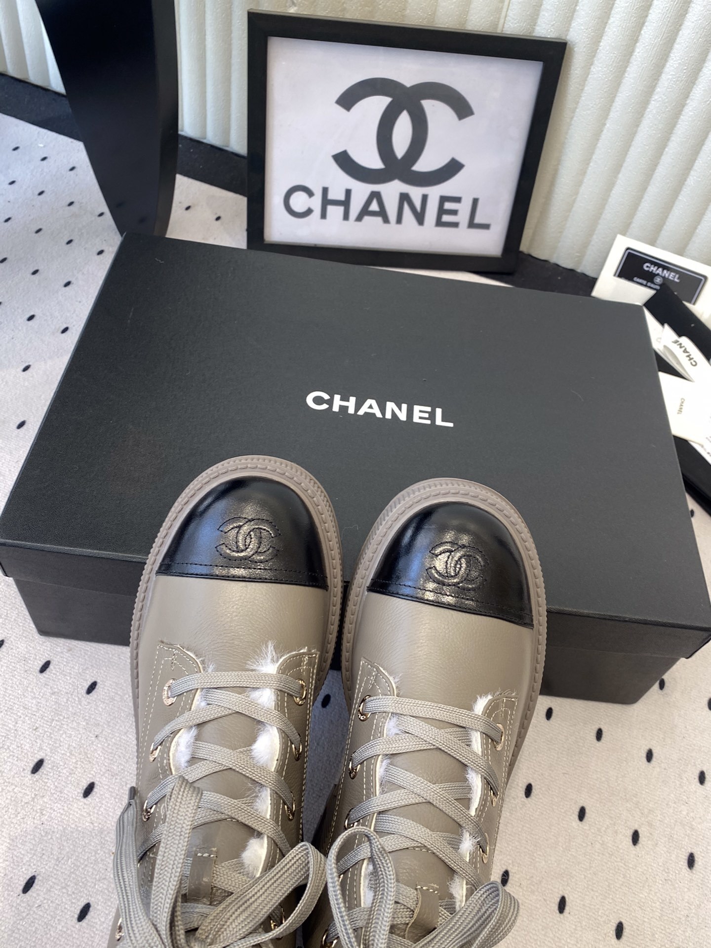 Chanel taupe leather lace-up boots with black patent toe caps and white shearling lining displayed on a black designer box - Hình 4