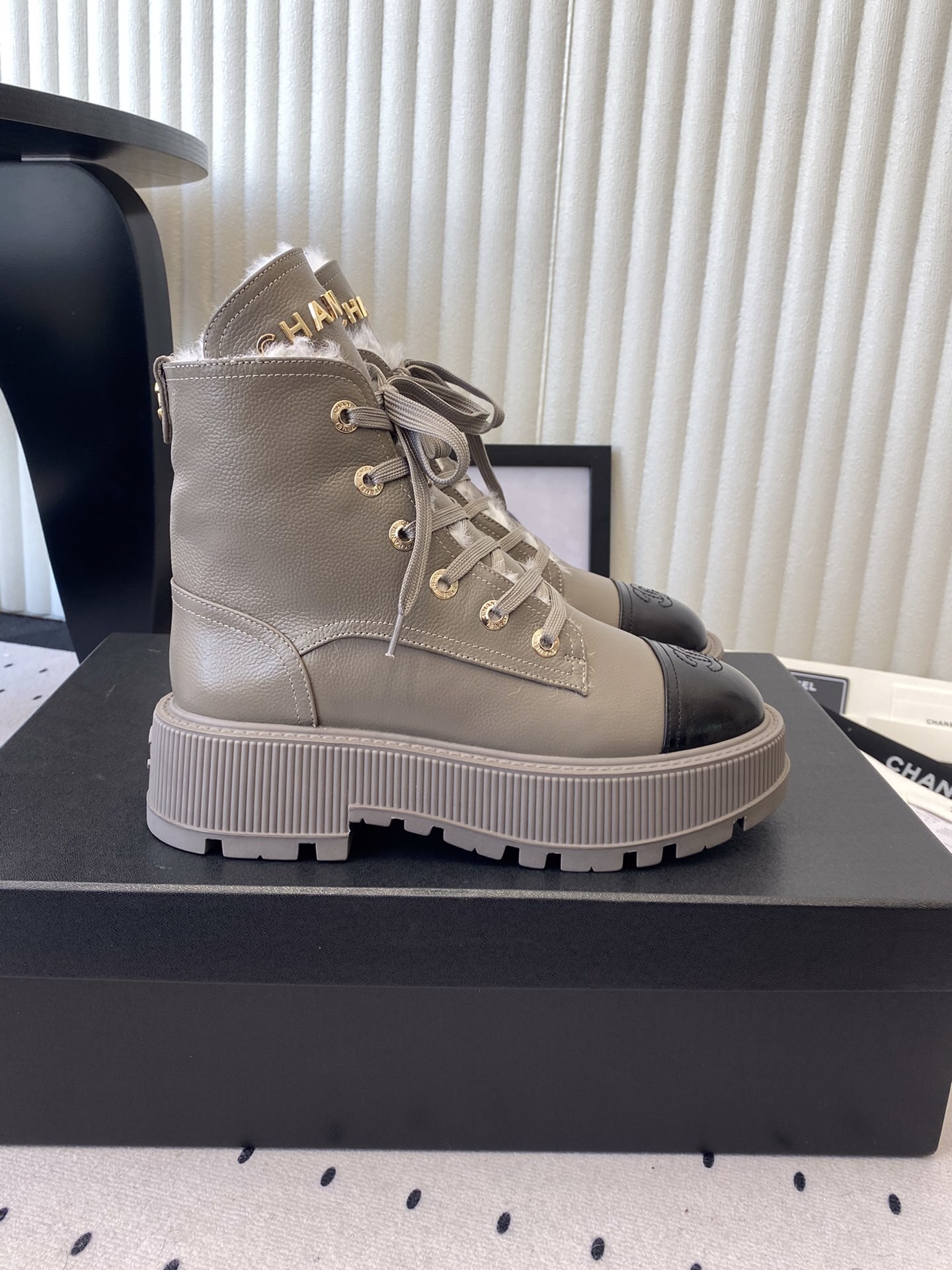 Chanel taupe leather lace-up boots with black patent toe caps and white shearling lining displayed on a black designer box - Hình 2