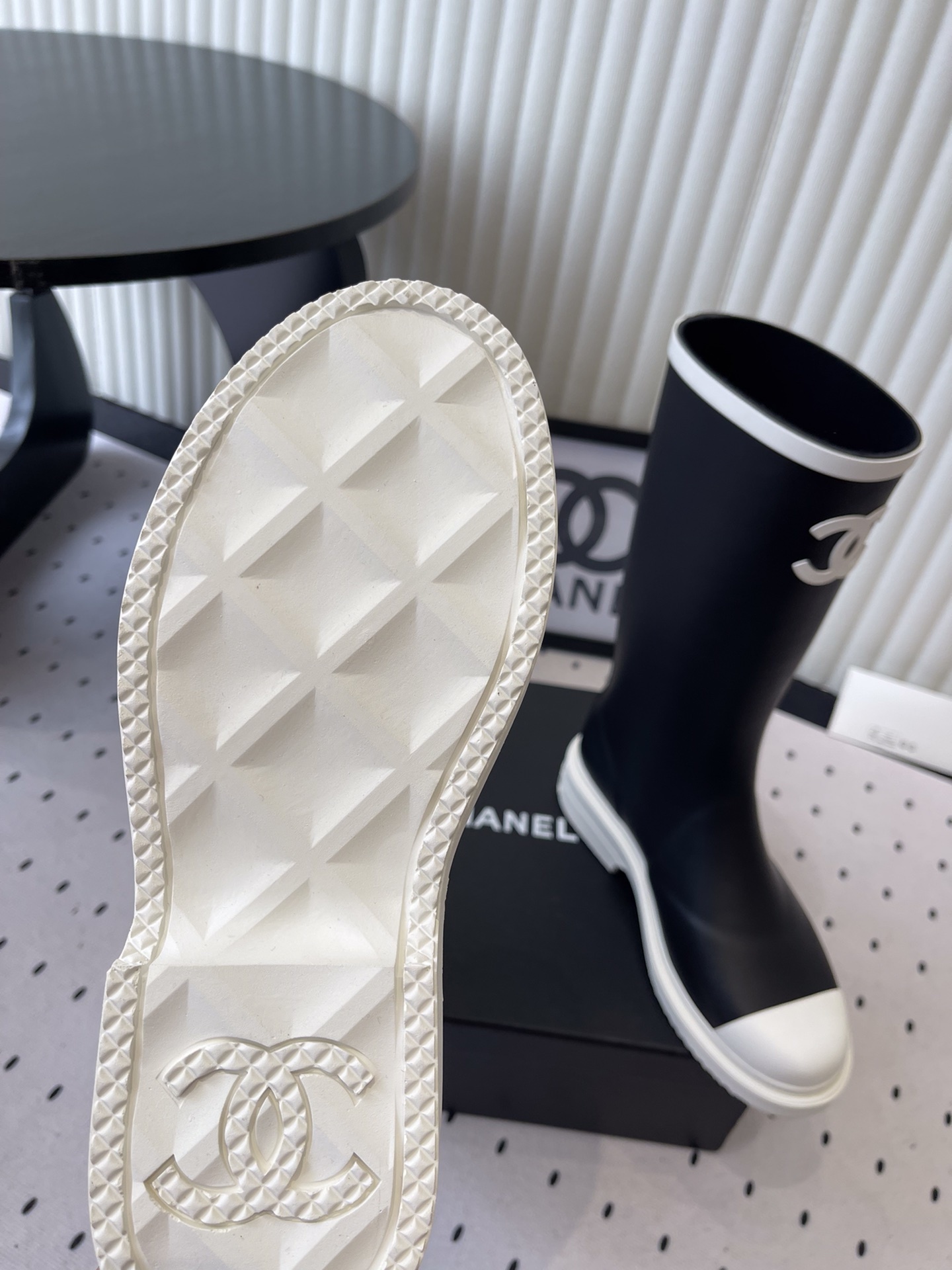 A pair of black Chanel rubber rain boots with white CC logo on the front and white soles, placed on a black Chanel shoe box. - Hình 9