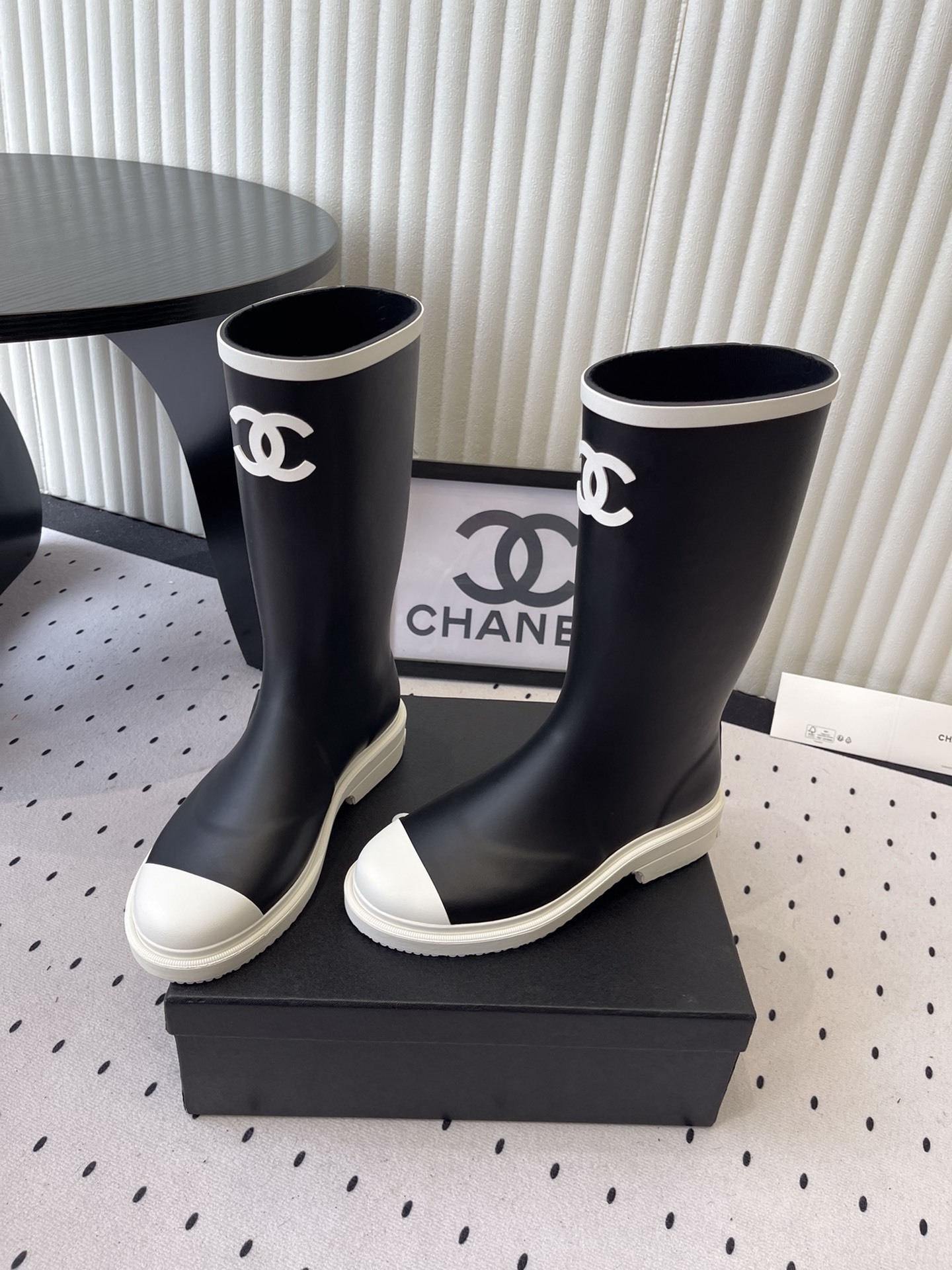 A pair of black Chanel rubber rain boots with white CC logo on the front and white soles, placed on a black Chanel shoe box. - Hình 8