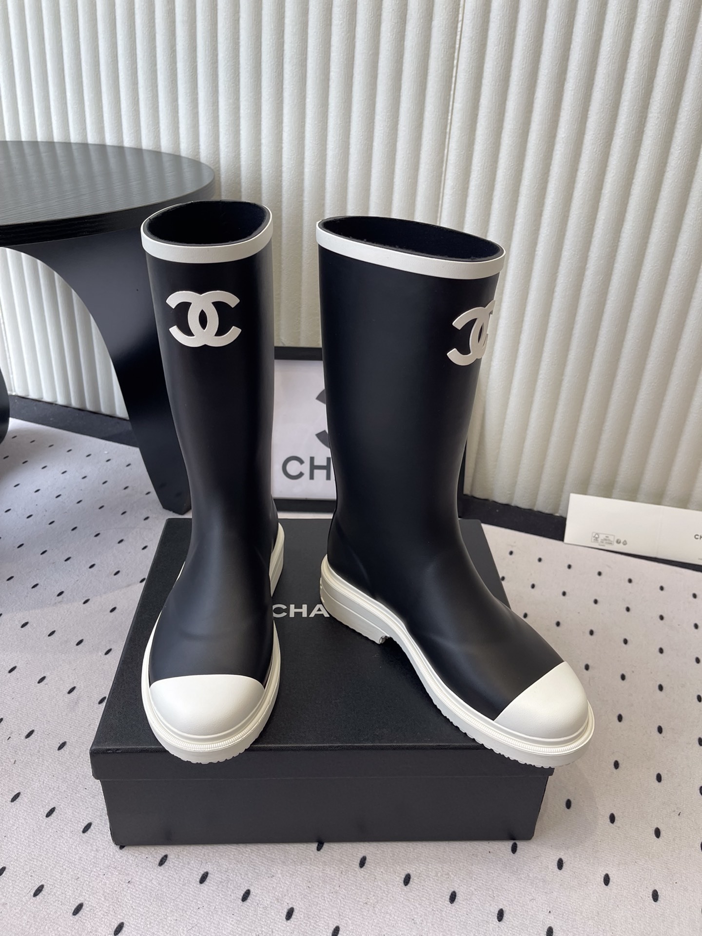 A pair of black Chanel rubber rain boots with white CC logo on the front and white soles, placed on a black Chanel shoe box. - Hình 7