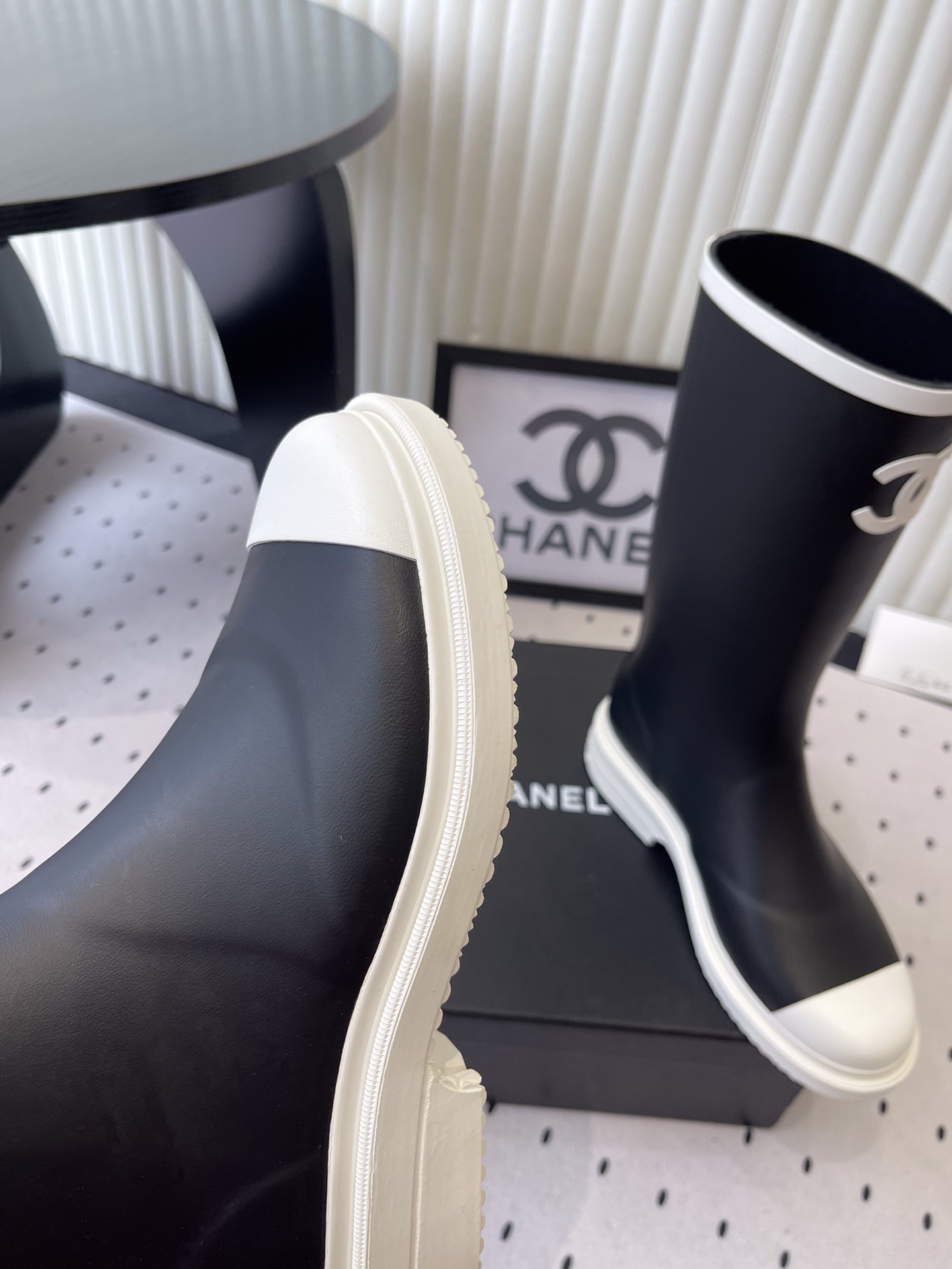 A pair of black Chanel rubber rain boots with white CC logo on the front and white soles, placed on a black Chanel shoe box. - Hình 6