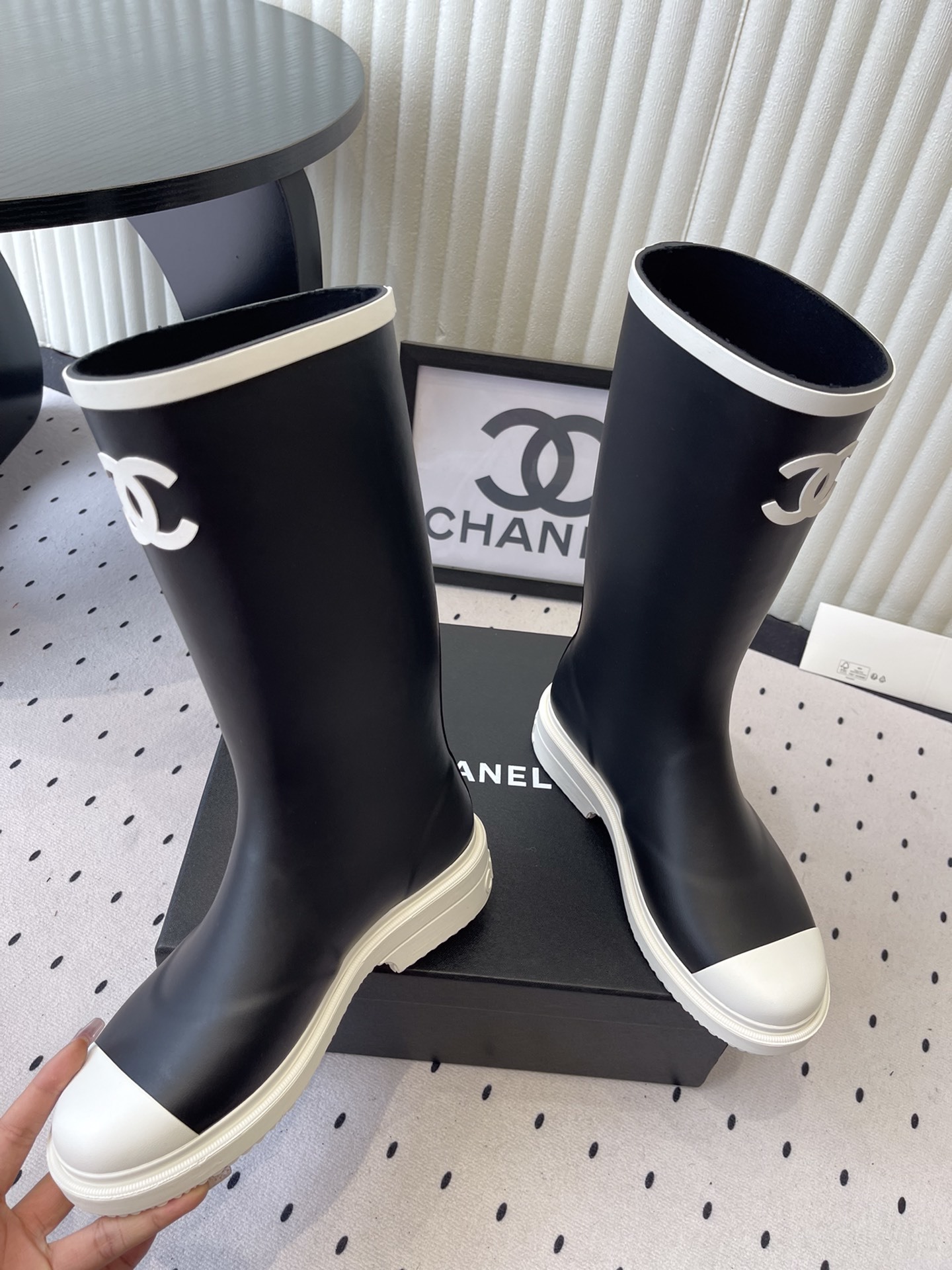 A pair of black Chanel rubber rain boots with white CC logo on the front and white soles, placed on a black Chanel shoe box. - Hình 5