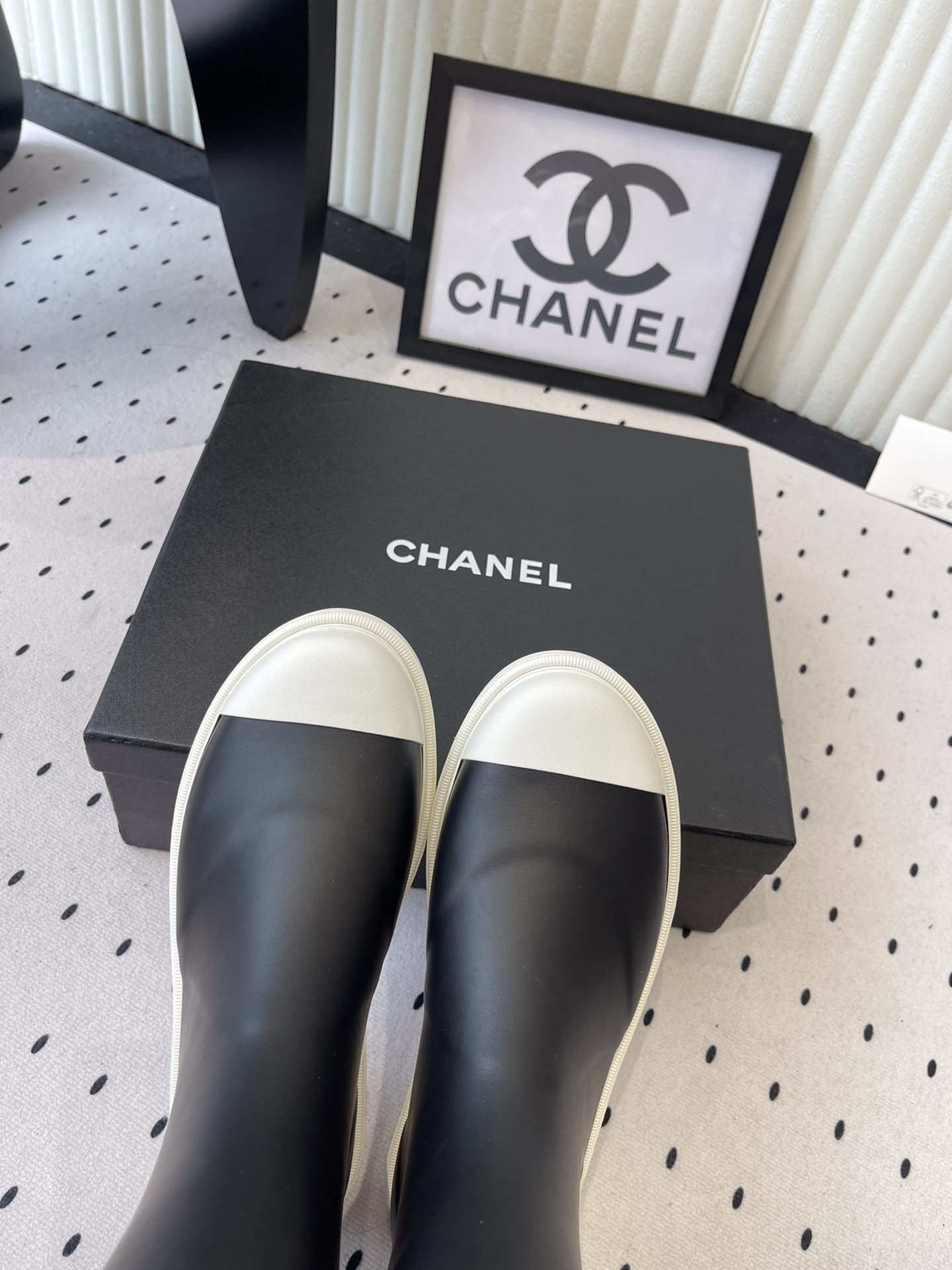 A pair of black Chanel rubber rain boots with white CC logo on the front and white soles, placed on a black Chanel shoe box. - Hình 4