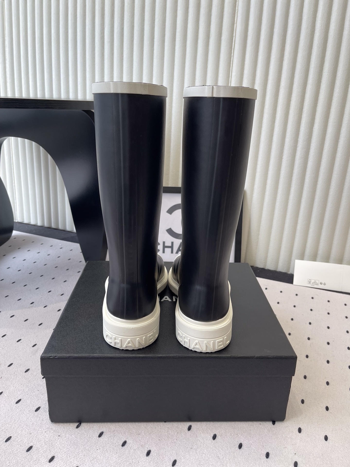 A pair of black Chanel rubber rain boots with white CC logo on the front and white soles, placed on a black Chanel shoe box. - Hình 3