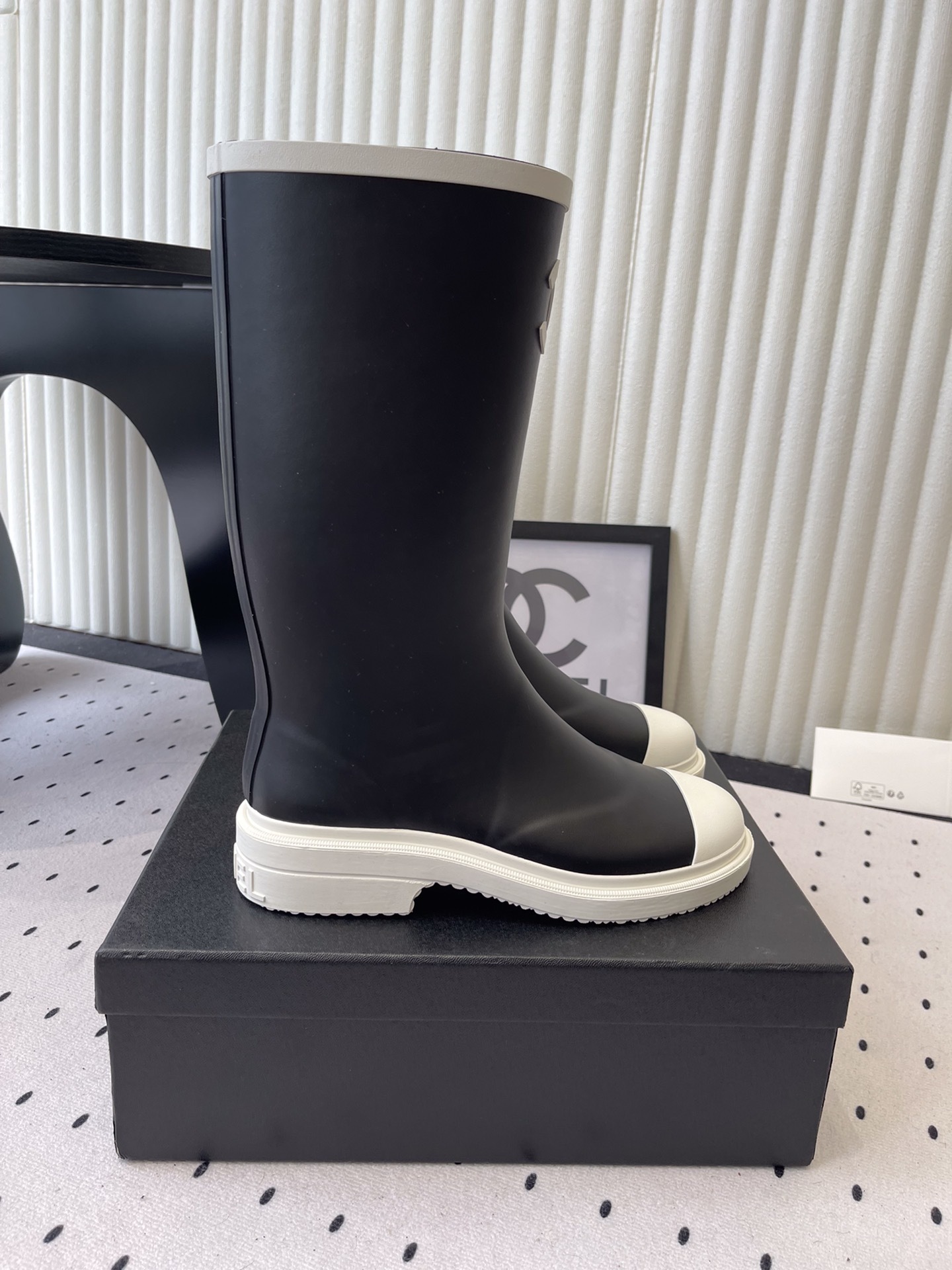 A pair of black Chanel rubber rain boots with white CC logo on the front and white soles, placed on a black Chanel shoe box. - Hình 2