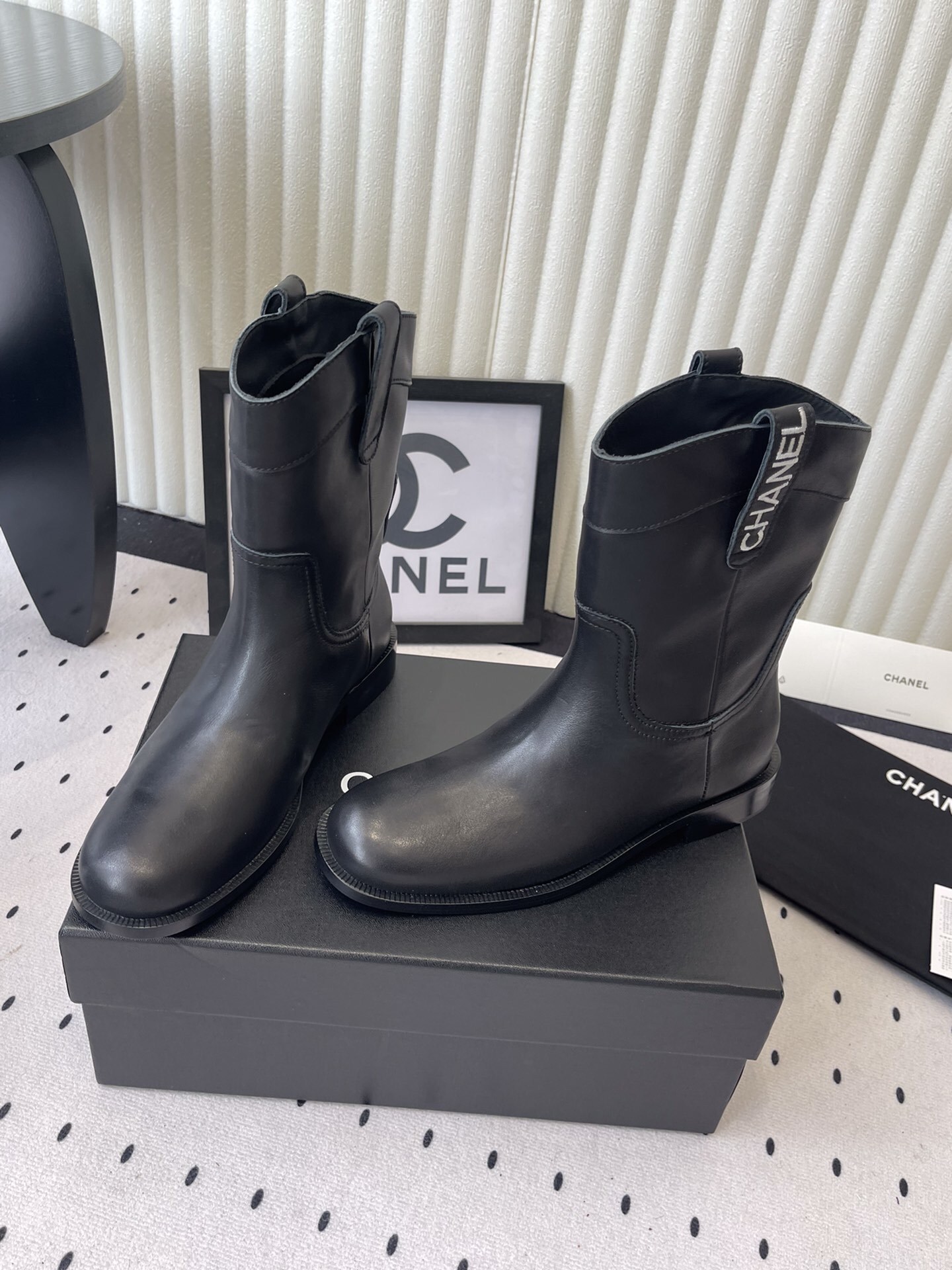 A pair of Chanel black leather mid-calf biker boots with white logo pull tabs displayed on a black Chanel shoe box. - Hình 8