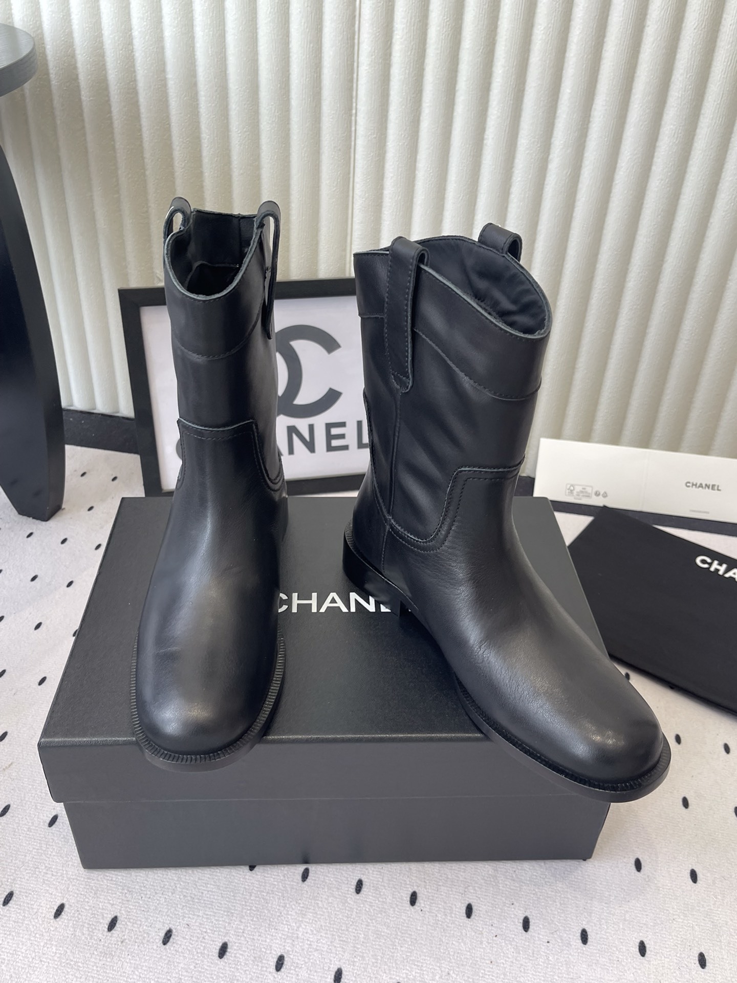 A pair of Chanel black leather mid-calf biker boots with white logo pull tabs displayed on a black Chanel shoe box. - Hình 7
