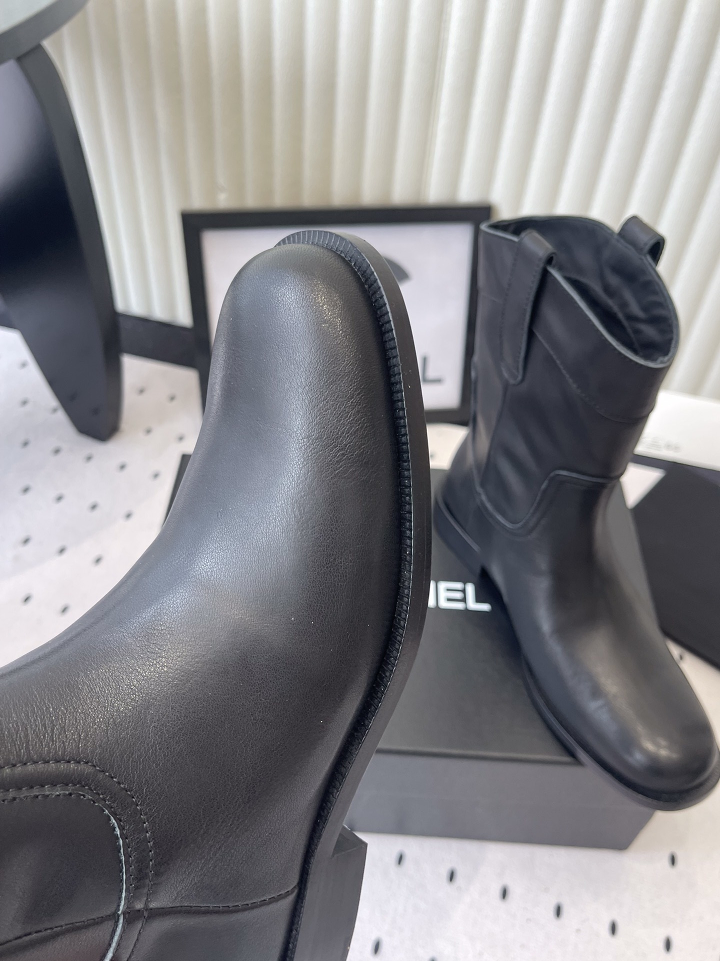 A pair of Chanel black leather mid-calf biker boots with white logo pull tabs displayed on a black Chanel shoe box. - Hình 6