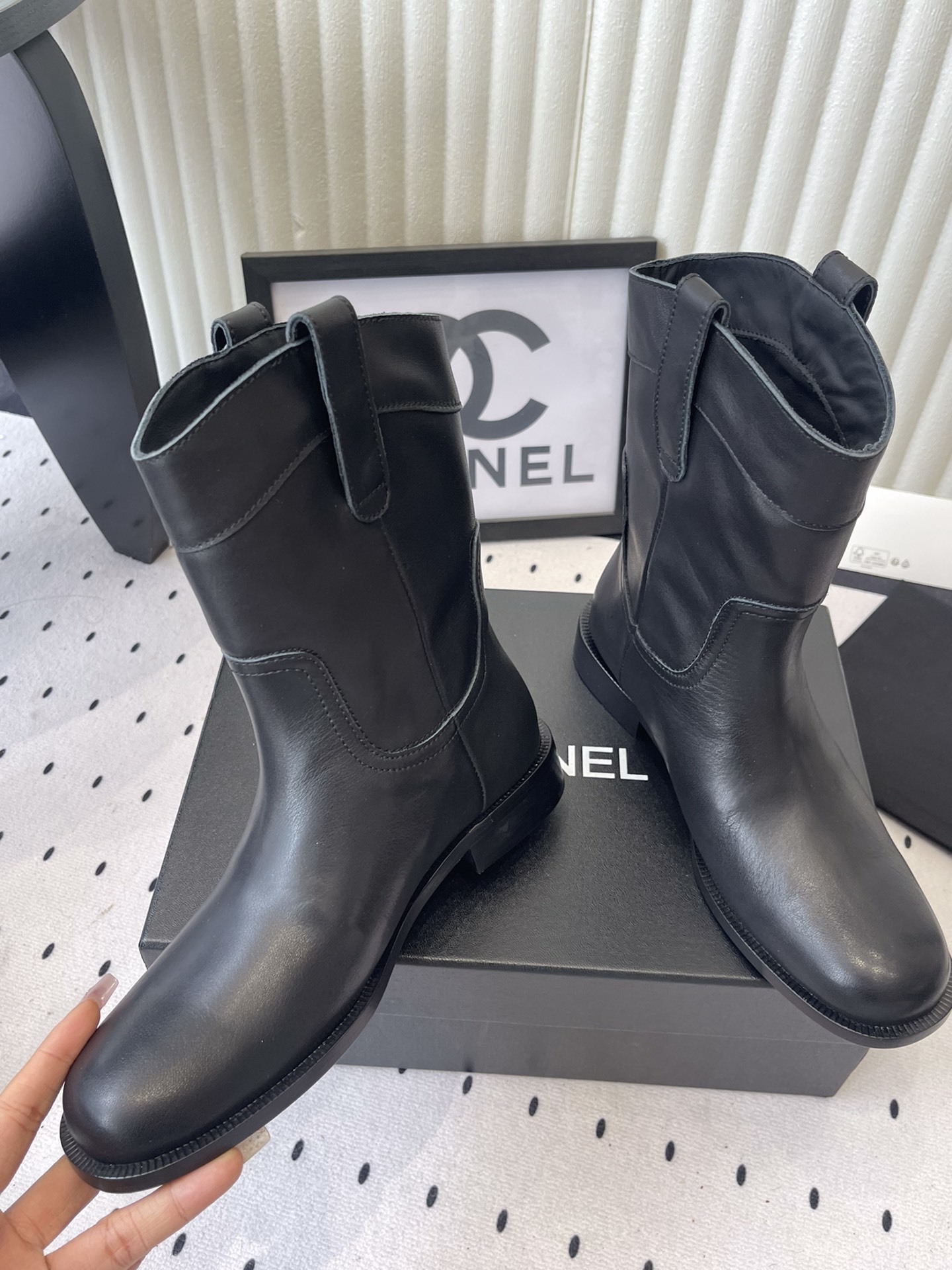 A pair of Chanel black leather mid-calf biker boots with white logo pull tabs displayed on a black Chanel shoe box. - Hình 5