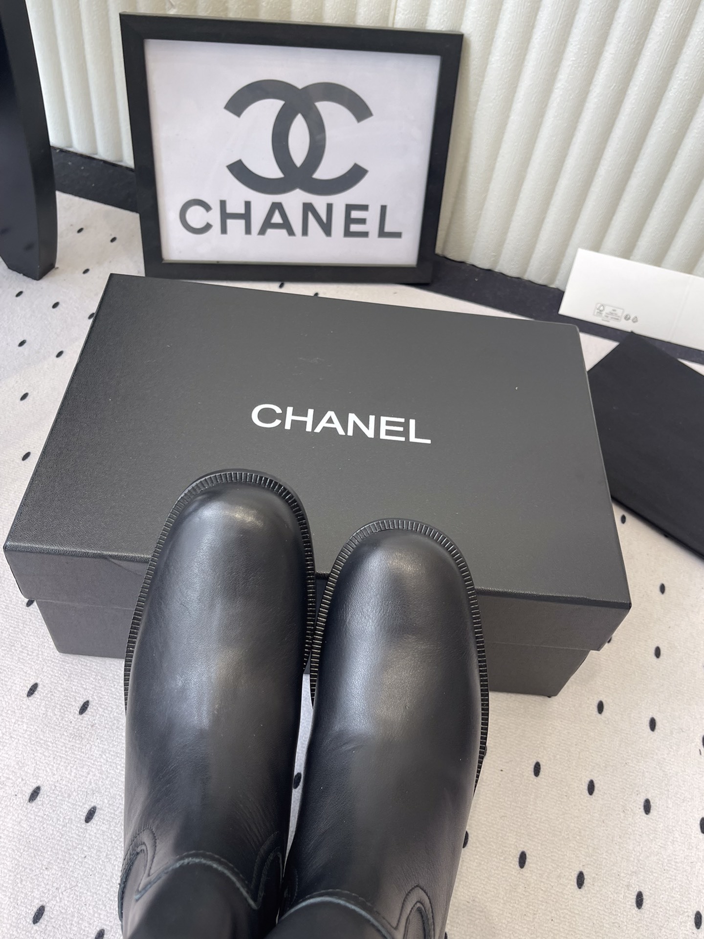 A pair of Chanel black leather mid-calf biker boots with white logo pull tabs displayed on a black Chanel shoe box. - Hình 4