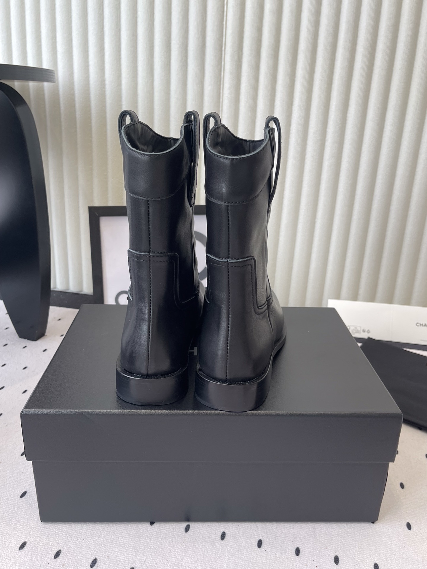 A pair of Chanel black leather mid-calf biker boots with white logo pull tabs displayed on a black Chanel shoe box. - Hình 3