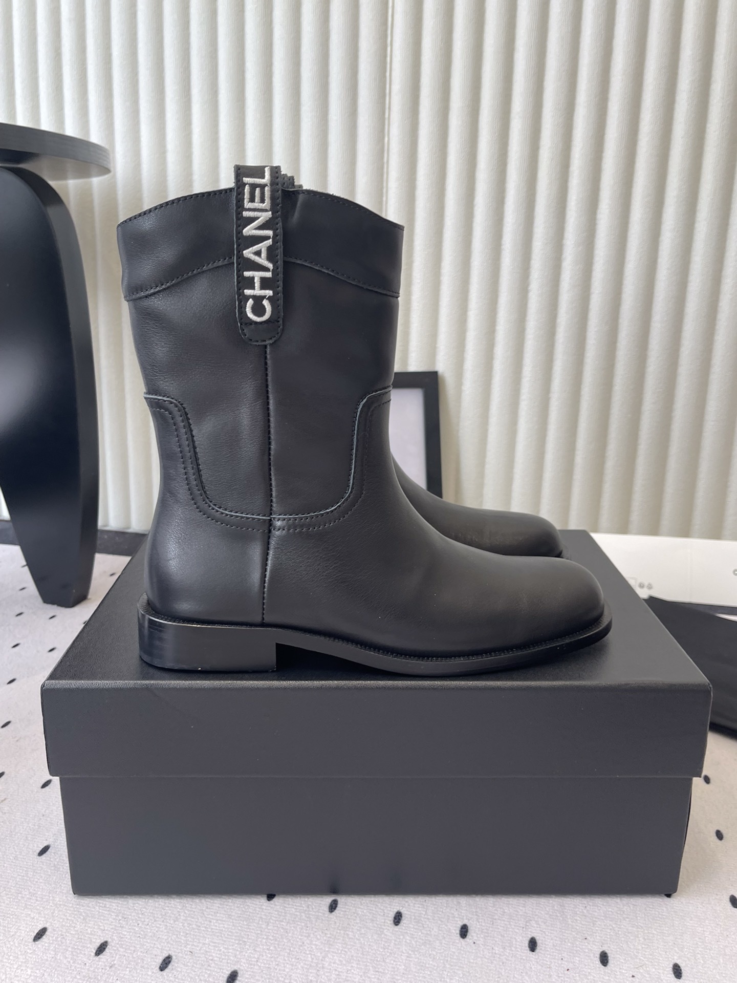 A pair of Chanel black leather mid-calf biker boots with white logo pull tabs displayed on a black Chanel shoe box. - Hình 2