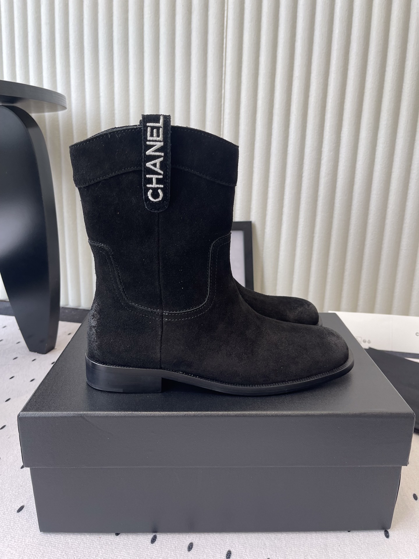 Pair of Chanel black suede mid-calf boots with white logo pull tabs displayed on a black Chanel shoe box - Hình 2
