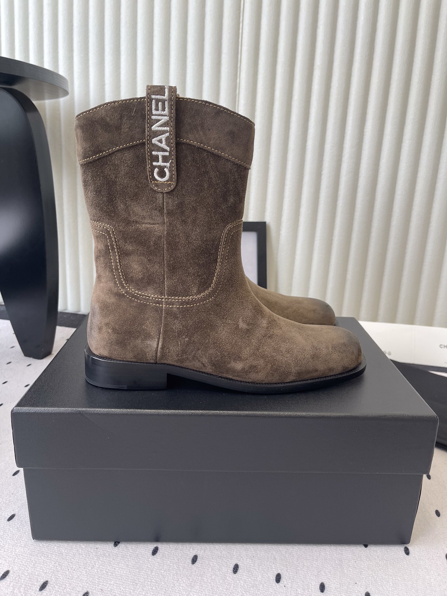 A pair of Chanel brown suede mid-calf Western style boots with embroidered logo pull tabs on a black box. - Hình 2