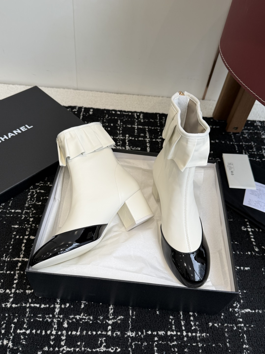 A pair of Chanel white leather ankle boots with black patent leather toe caps and ruffled collars on a black designer box. - Hình 5