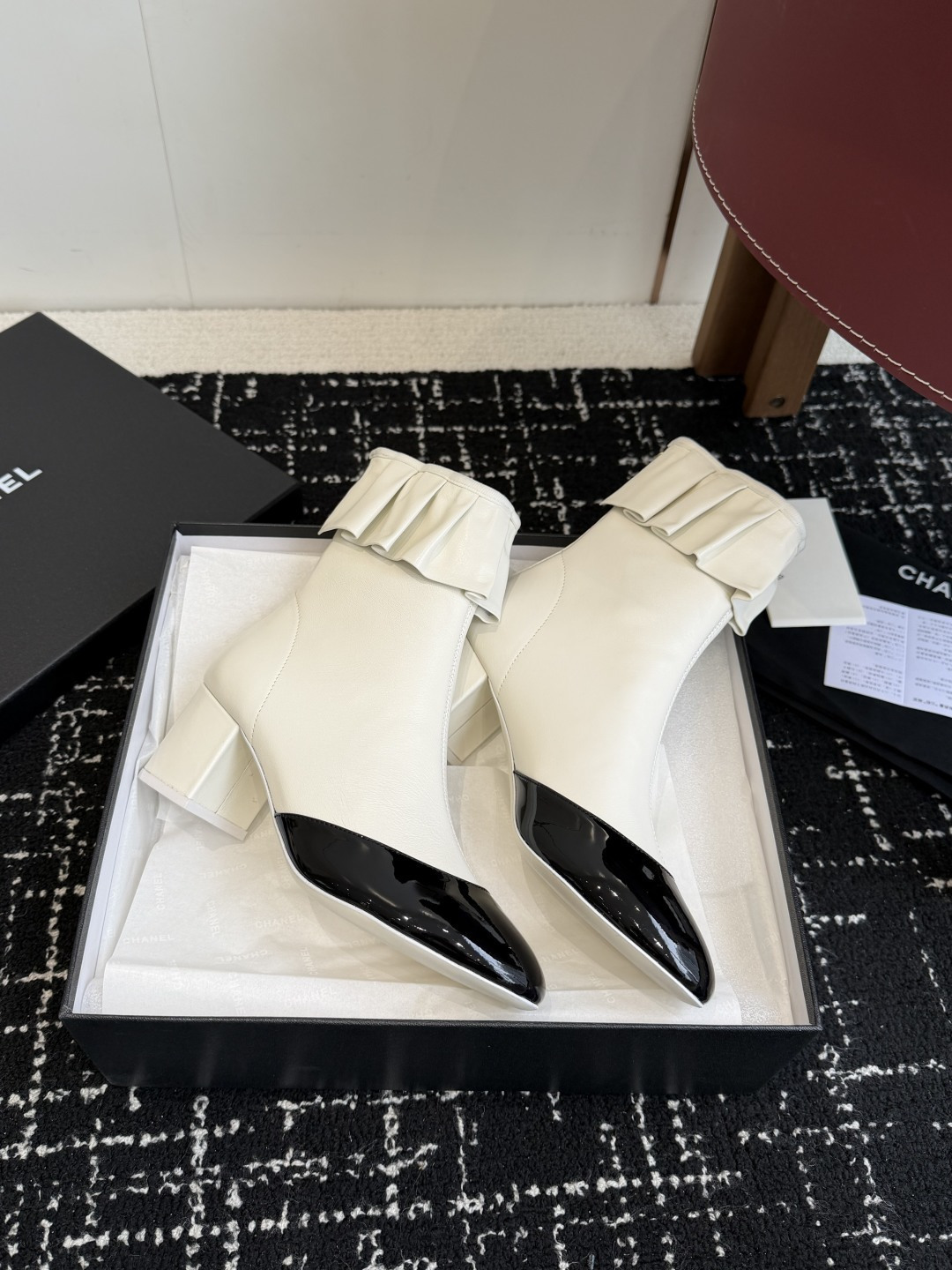 A pair of Chanel white leather ankle boots with black patent leather toe caps and ruffled collars on a black designer box. - Hình 4