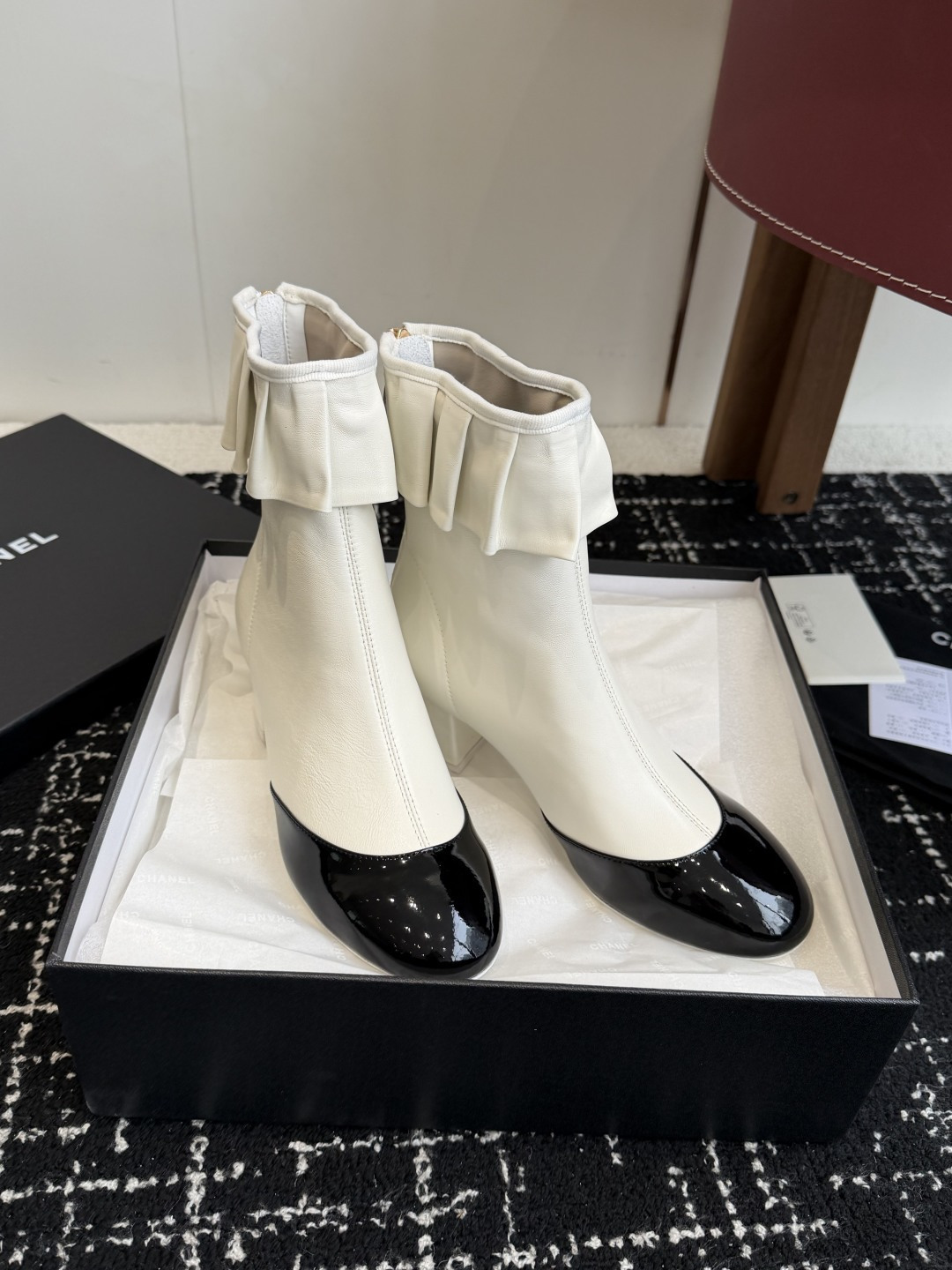 A pair of Chanel white leather ankle boots with black patent leather toe caps and ruffled collars on a black designer box. - Hình 3