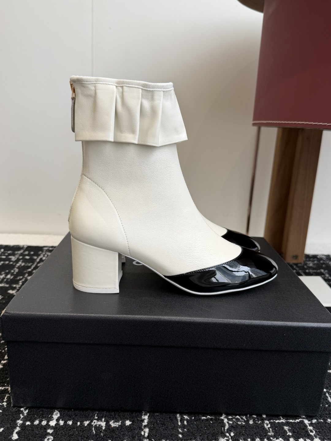 A pair of Chanel white leather ankle boots with black patent leather toe caps and ruffled collars on a black designer box. - Hình 2