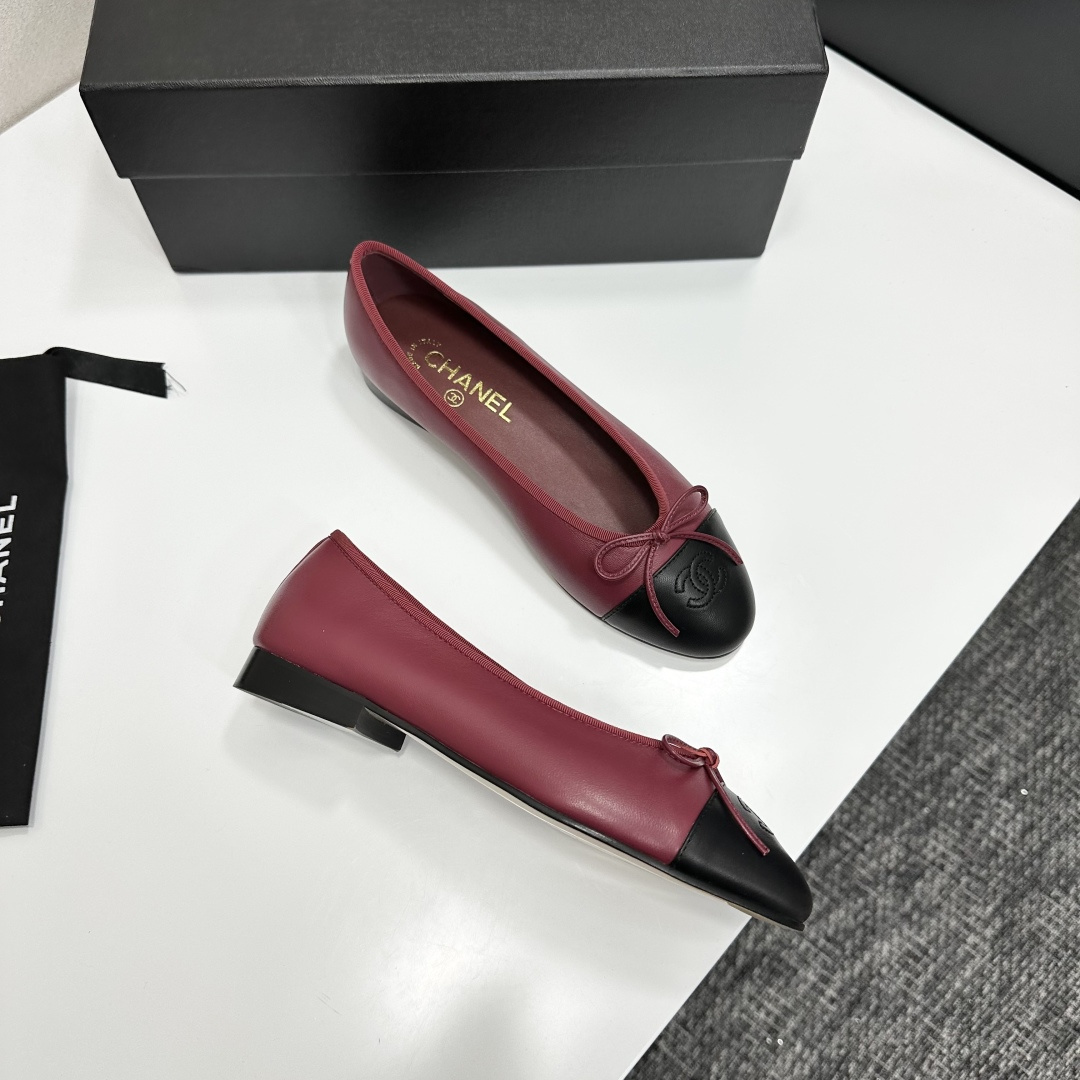 A pair of burgundy leather ballet flats with black cap toes and bow detail resting on a black designer box. - Hình 10