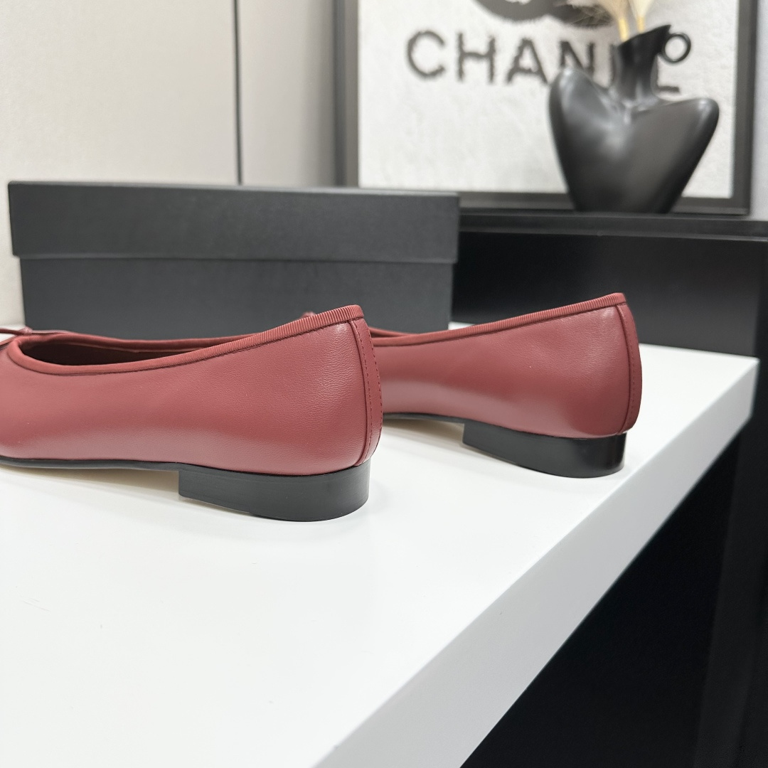 A pair of burgundy leather ballet flats with black cap toes and bow detail resting on a black designer box. - Hình 9