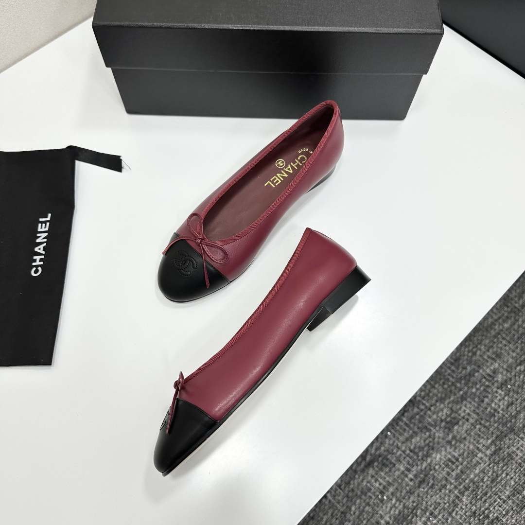 A pair of burgundy leather ballet flats with black cap toes and bow detail resting on a black designer box. - Hình 7