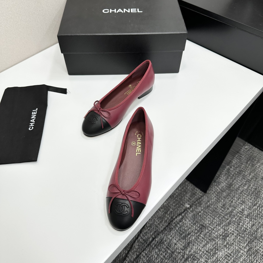 A pair of burgundy leather ballet flats with black cap toes and bow detail resting on a black designer box. - Hình 6