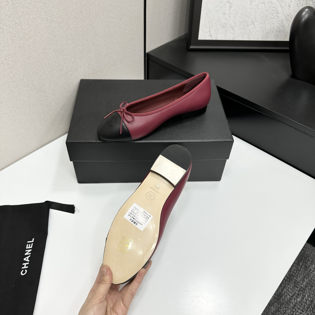 A pair of burgundy leather ballet flats with black cap toes and bow detail resting on a black designer box. - Hình 5