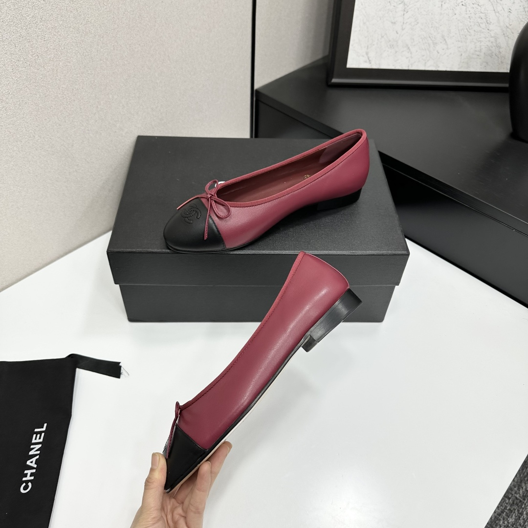 A pair of burgundy leather ballet flats with black cap toes and bow detail resting on a black designer box. - Hình 4