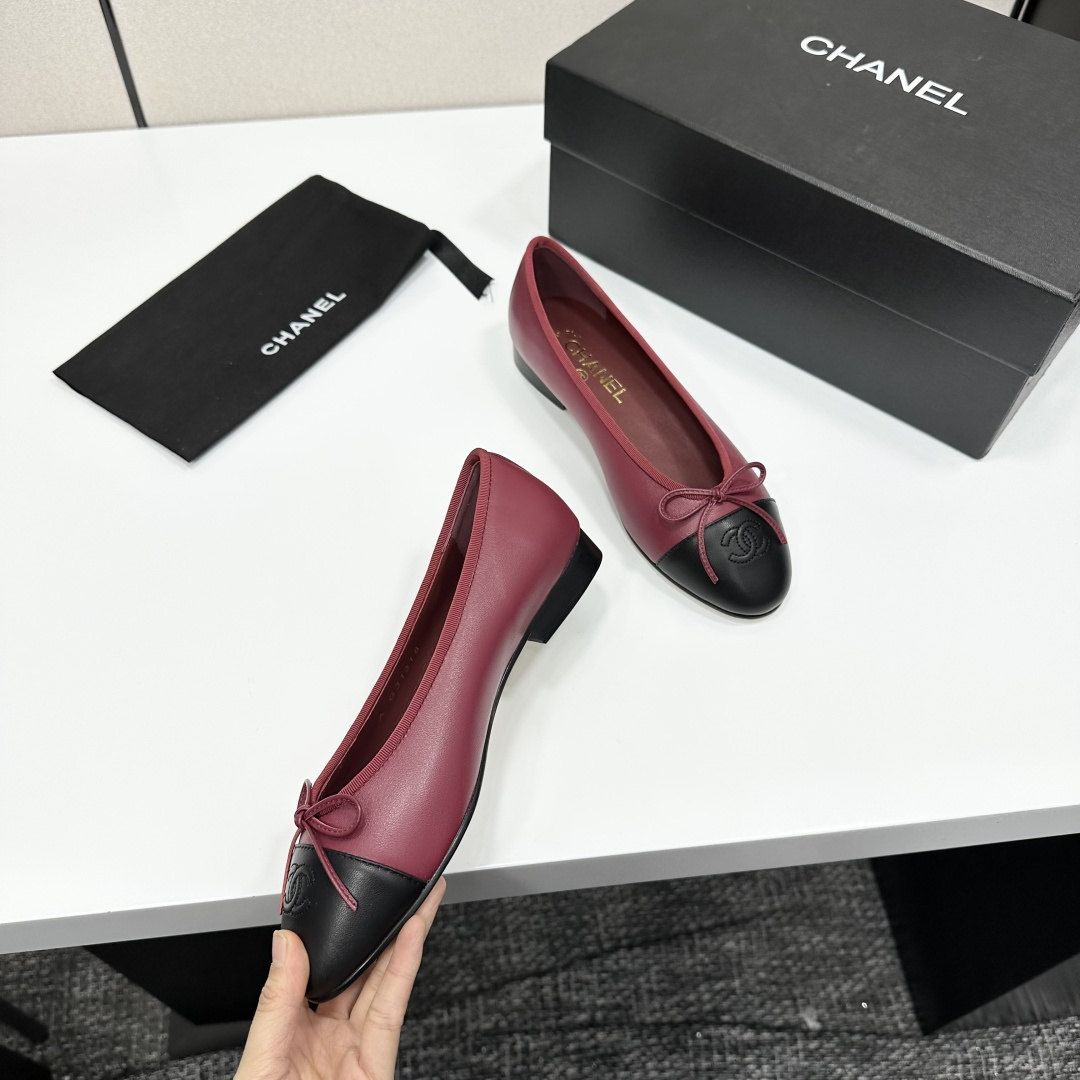 A pair of burgundy leather ballet flats with black cap toes and bow detail resting on a black designer box. - Hình 2