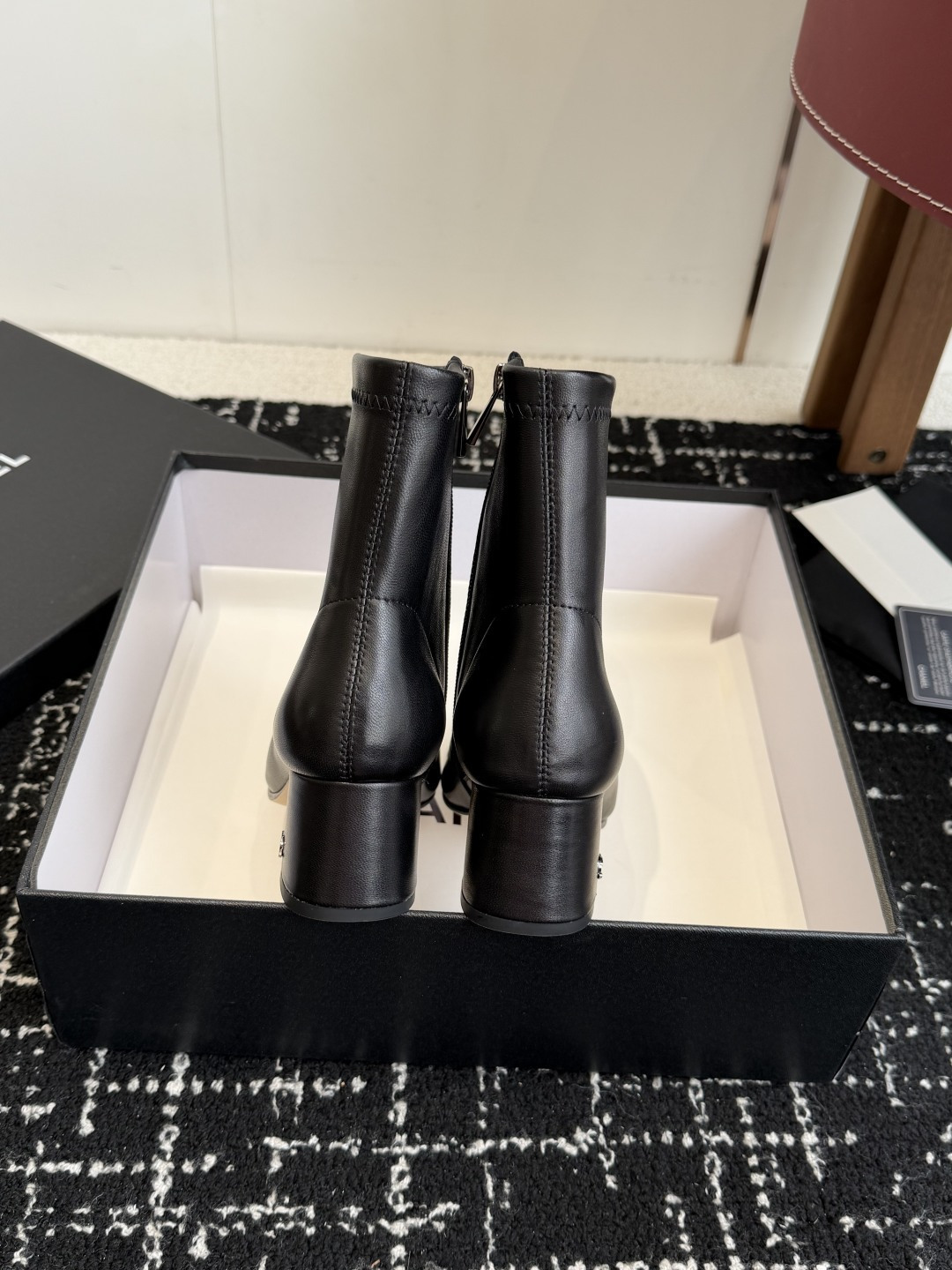 A pair of black Chanel leather ankle boots with glossy patent leather toe caps and block heels featuring the CC logo, displayed on a black box. - Hình 8