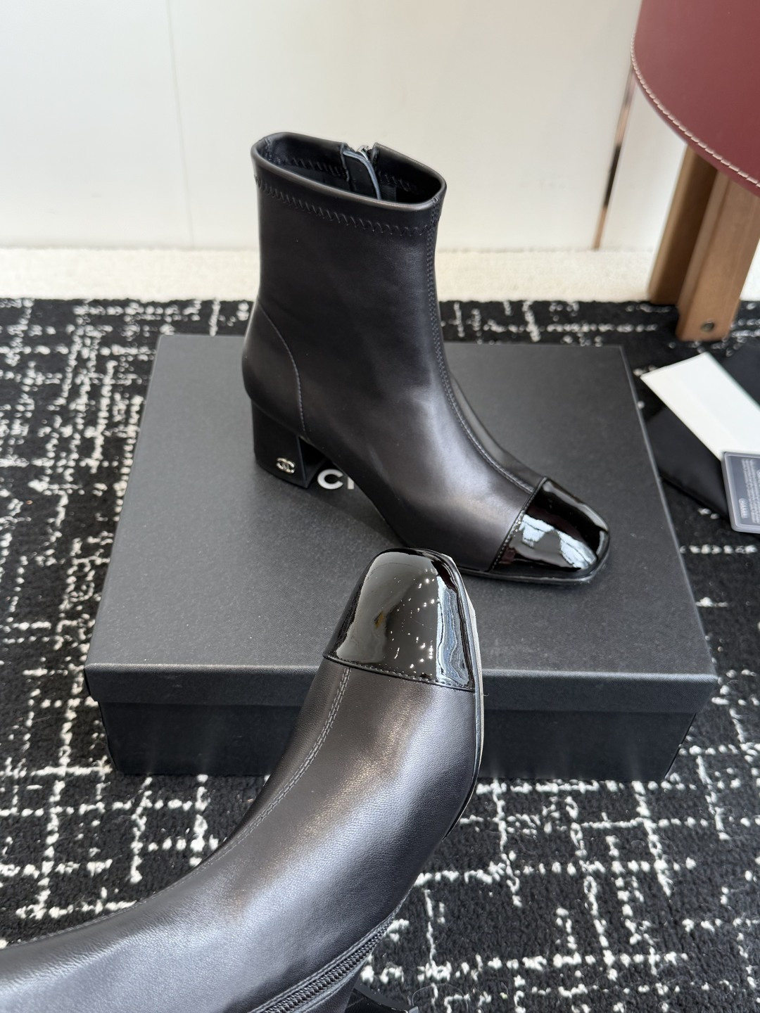 A pair of black Chanel leather ankle boots with glossy patent leather toe caps and block heels featuring the CC logo, displayed on a black box. - Hình 7