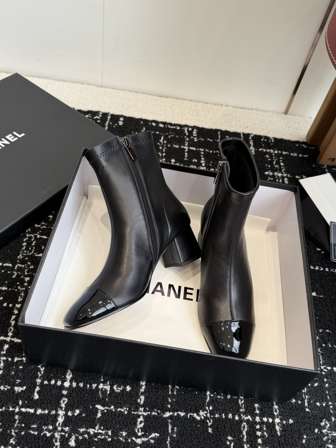 A pair of black Chanel leather ankle boots with glossy patent leather toe caps and block heels featuring the CC logo, displayed on a black box. - Hình 6