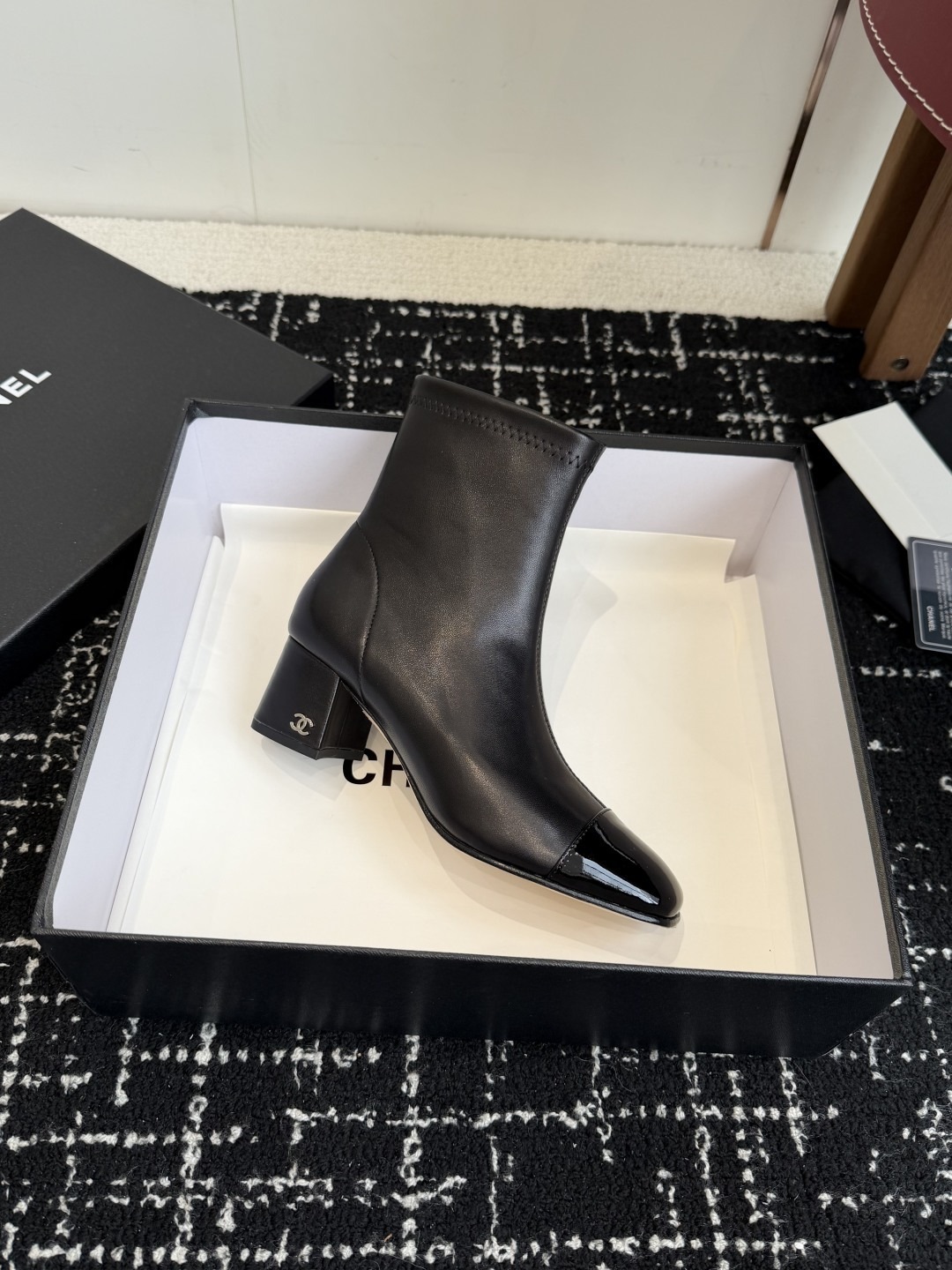 A pair of black Chanel leather ankle boots with glossy patent leather toe caps and block heels featuring the CC logo, displayed on a black box. - Hình 5