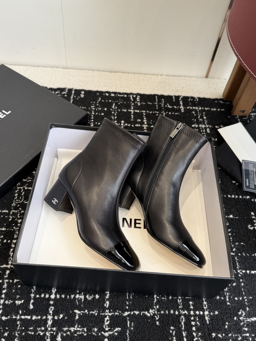 A pair of black Chanel leather ankle boots with glossy patent leather toe caps and block heels featuring the CC logo, displayed on a black box. - Hình 4