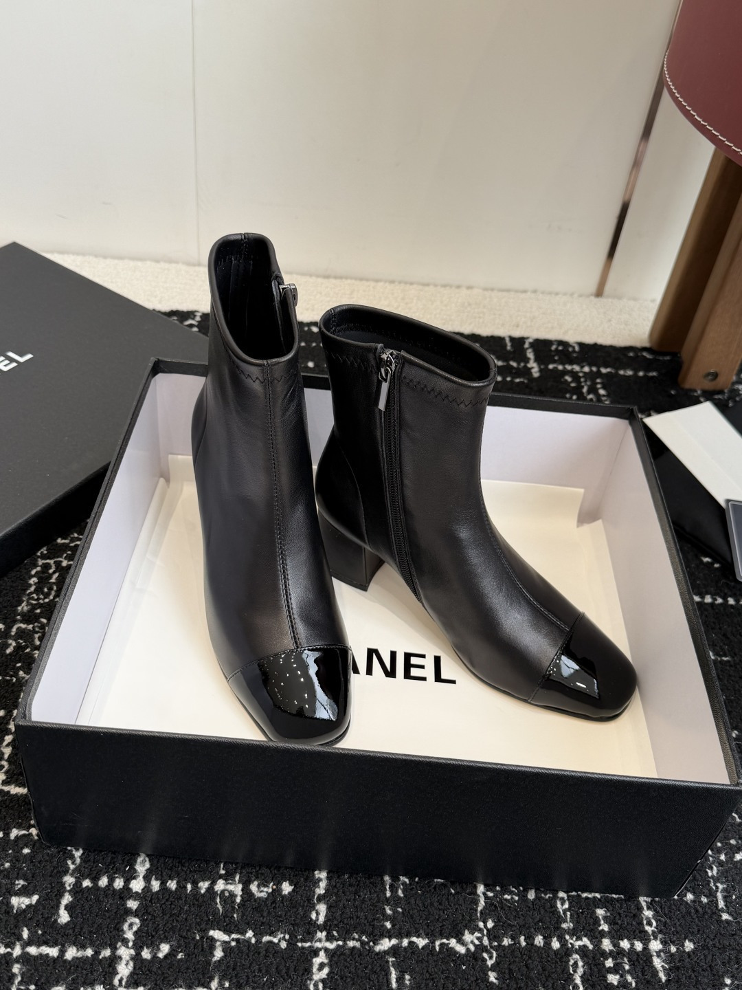 A pair of black Chanel leather ankle boots with glossy patent leather toe caps and block heels featuring the CC logo, displayed on a black box. - Hình 3